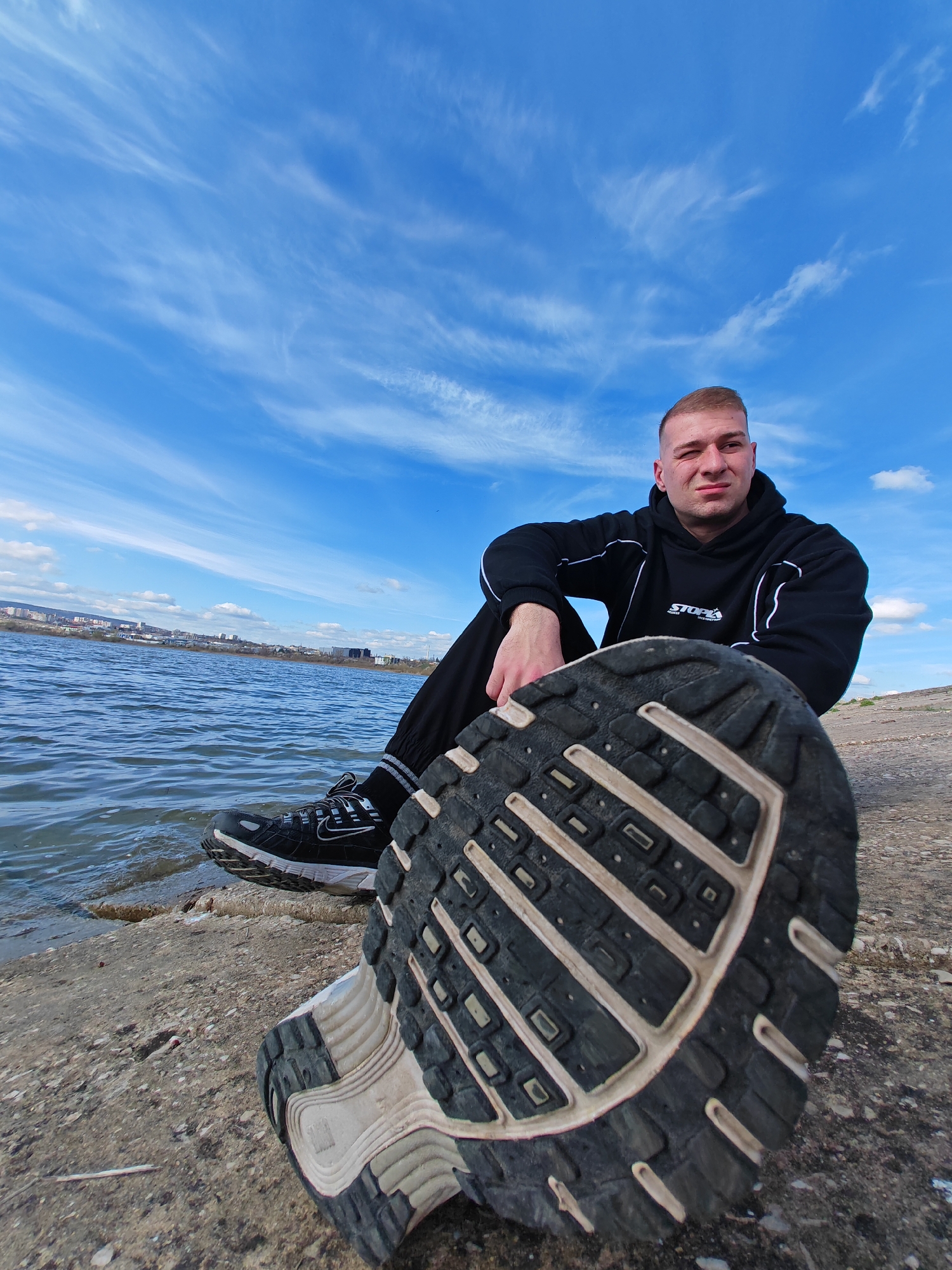 A medium-shot depicts a man sitting on a concrete bank next to a body of water. The man is fair-skinned, with short-cropped hair, and is wearing a black tracksuit with white trim. He is looking directly at the camera. His right leg is bent, with his foot planted on the ground. In the foreground, the sole of a running shoe is prominently displayed, obscuring part of the scene. The edge of the water is visible to the left, and a cityscape is visible on the opposite side of the water. The sky above is blue with wispy clouds. The location is near Shumen, Bulgaria, identified by the cityscape across the lake.
