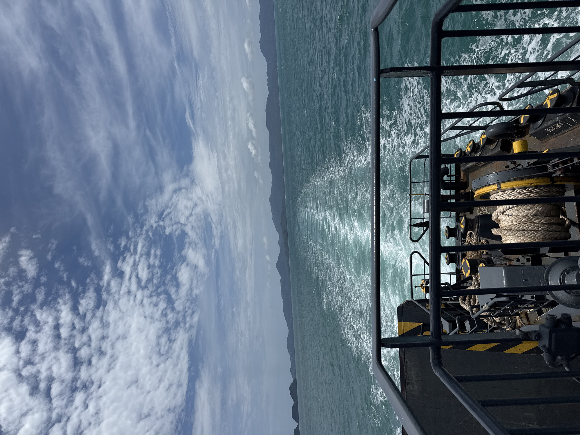 An image taken from the rear of a ferry on a bright, cloudy day. The ferry is moving forward, creating a white wake in the turquoise water. In the distance, a chain of lush green islands with mountainous terrain is visible against a pale blue sky dotted with white, wispy clouds. The foreground of the image shows the deck of the ferry with metal railings, ropes, and winches. Yellow and black hazard stripes are painted on some of the ferry's structures. There are no people visible in the image. The scene depicts travel by sea between islands, likely in the vicinity of Ko Pha-ngan, Thailand, given the tropical island landscape.
