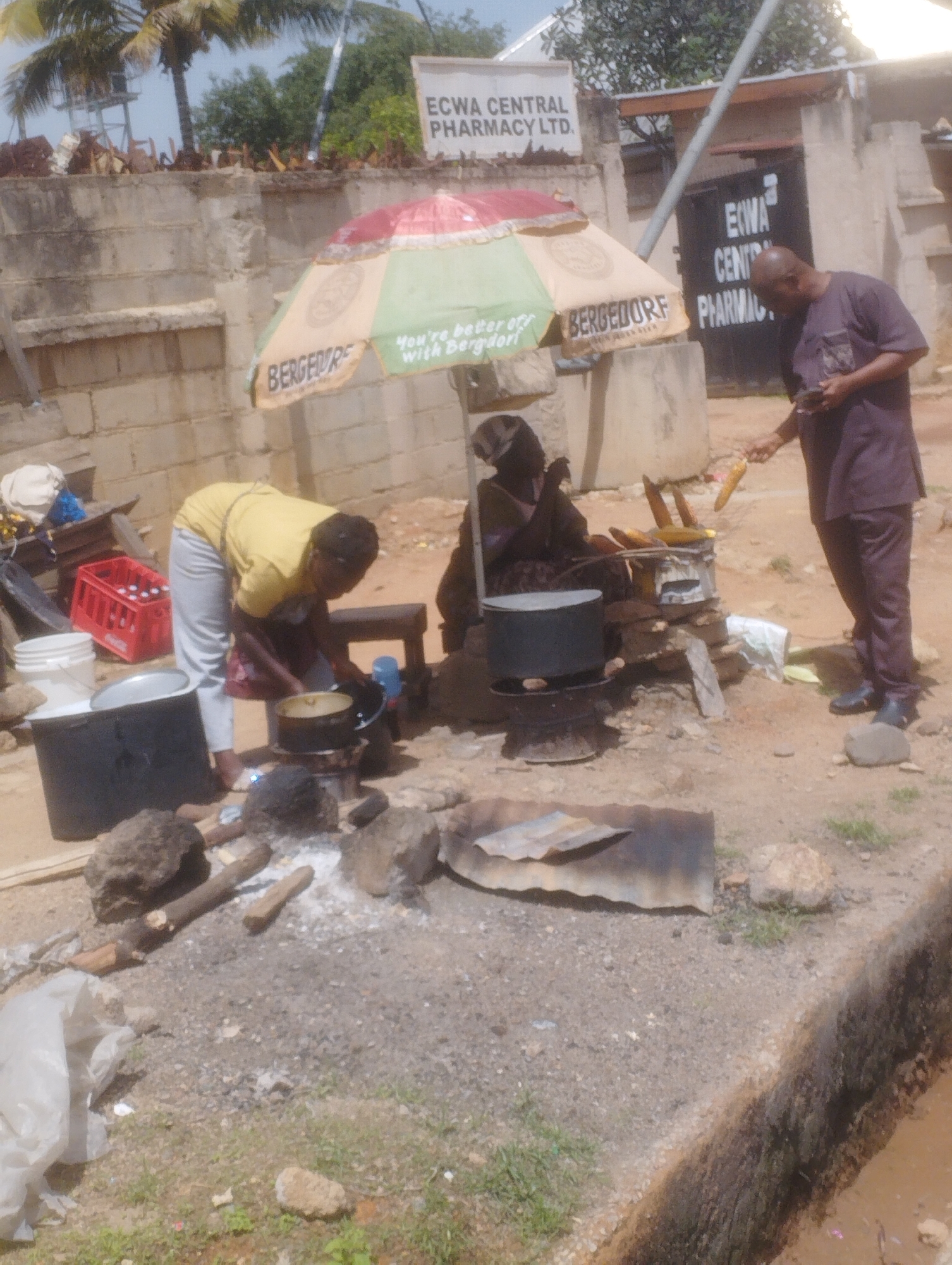 In Jos, Nigeria, a street vendor prepares corn for sale. The scene depicts an outdoor market stall with a woman wearing a yellow shirt and grey pants, bent over a cooking pot. Adjacent to her, a man in a dark brown traditional outfit is holding a piece of corn, seemingly inspecting it or waiting to purchase it. The stall features a colorful umbrella offering shade, with the words "BERGEDORF" and "You're better with BERGEDORF" visible. Various cooking implements, including pots and what appear to be makeshift grills, are scattered around. In the background, a building with signage for "ECWA CENTRAL PHARMACY LTD." is visible, suggesting the location is near this establishment. The ground is dusty with some scattered debris, indicating a typical urban street setting in Jos.