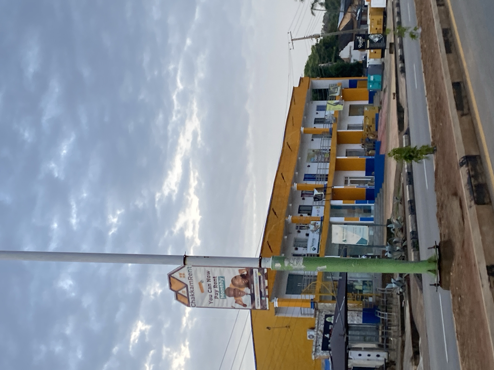 This is a medium-shot, eye-level, outdoor photograph of a street scene in Jos, Nigeria, taken on a cloudy day. The primary subject is a two-story commercial building painted in bright yellow and white, with blue accents on the lower level. The building has a row of columns on the upper level, supporting an overhang. Several businesses appear to be housed within the building, with signage visible. A prominent billboard on a pole in front of the building advertises "ChakkamRent" and states, "You Can Now Pay Rent Monthly."
In the foreground, a paved road runs horizontally across the frame, with a concrete divider and some sparse vegetation. The road is marked with white lines. The street also features a green utility pole in the middle ground. Various vehicles, including what appear to be generators or other heavy equipment, are parked in front of the building. The sky is heavily overcast with gray clouds. The overall impression is that of a typical urban commercial area in Jos.