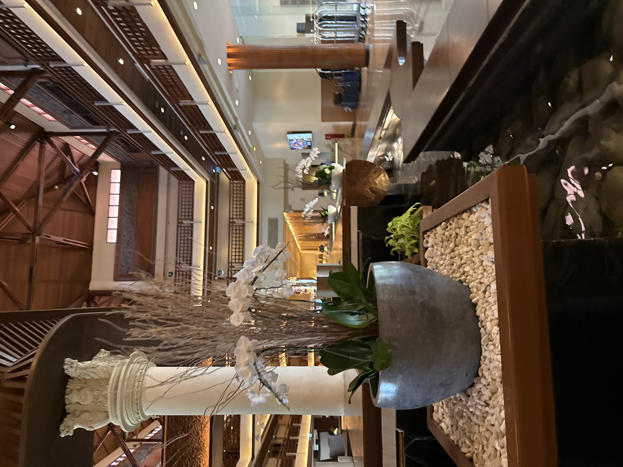 The image shows the interior of a building in Nusa Dua, Indonesia. The architecture features a blend of traditional and modern elements, with a high, wooden-beam ceiling and open structure, contrasted by contemporary lighting and design. There are ornamental features like a stone-based pillar, decorative plants in a pot, and a water feature edged with small white stones, creating a tranquil ambiance. The overall environment is elegant and likely within a hotel or resort. There are no people.