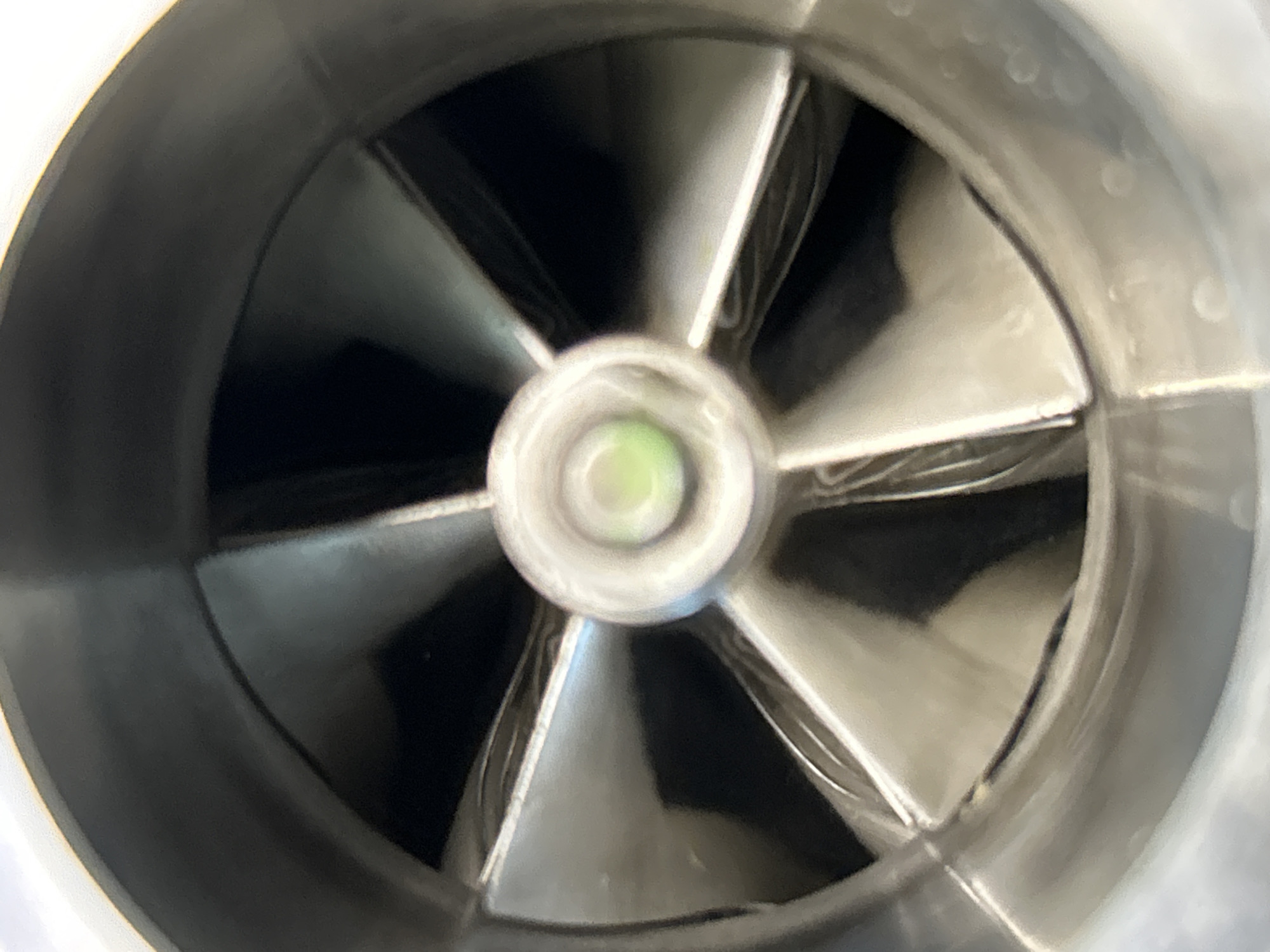 The image shows a close-up, head-on view of a fan or impeller with six blades. The blades are silver-colored and curved, and they converge towards a central hub. In the center of the hub is a small, circular, greenish-yellow light or reflective surface. The fan is housed within a metallic cylindrical casing, which is primarily silver and black. The lighting suggests it might be indoors, possibly in a workshop or industrial setting. There are no people or other discernable objects in the image. There is no information in the image to suggest a location in Tallinn, Estonia.
