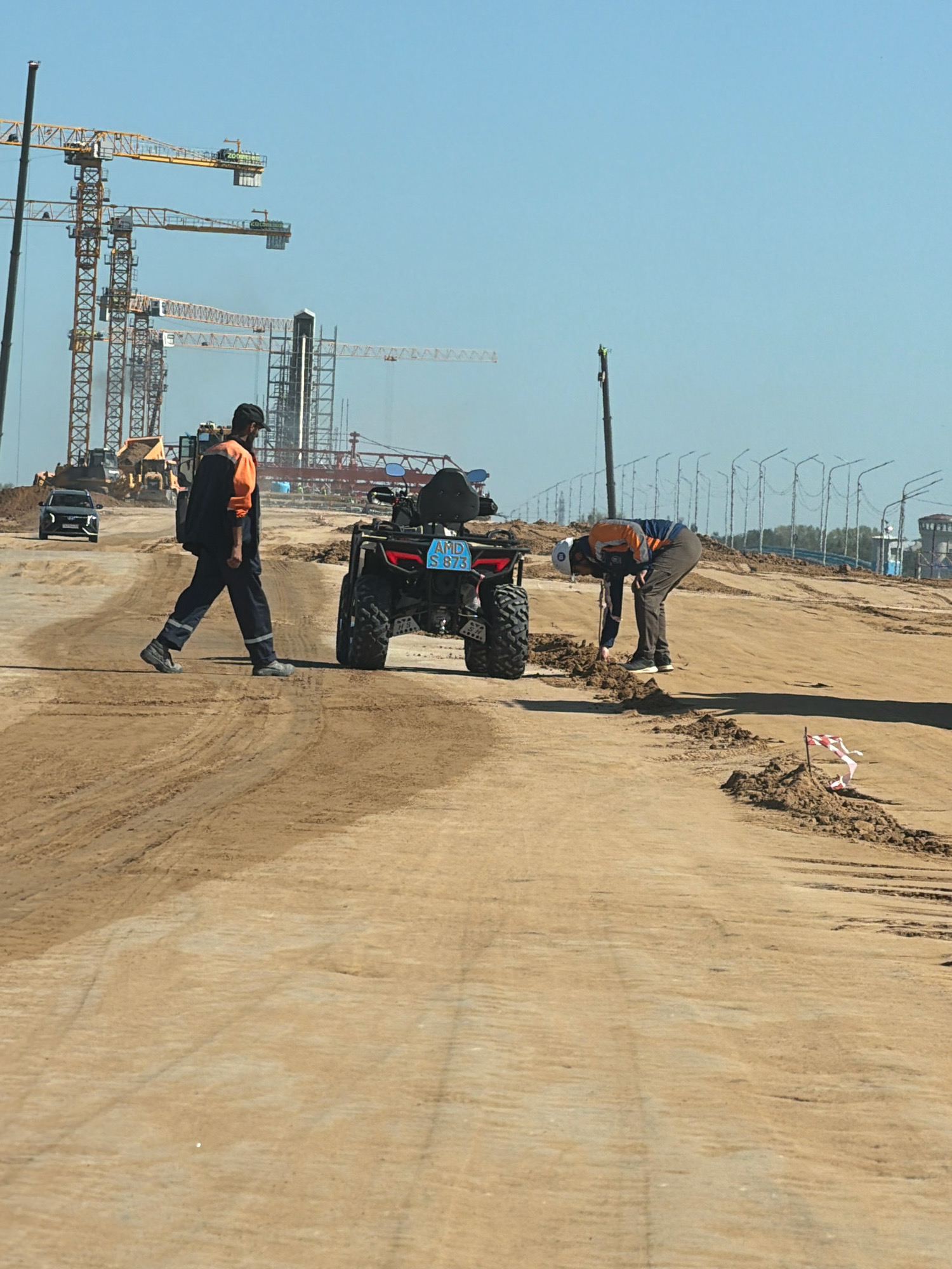 In the city of Efremovka, Kazakhstan, a construction site is depicted under a clear blue sky. Two men in work attire are present. One man, wearing an orange and navy work jacket, dark pants, and a black cap, walks across a dirt path. He is carrying a dark bag over his shoulder. Beside him is a black all-terrain vehicle (ATV) with a blue license plate reading "AMD S 873". The second man, wearing a white helmet, an orange and navy work jacket, and dark pants, is bending down next to the ATV, appearing to examine the ground. The background shows numerous construction cranes and structures, indicating ongoing development. The ground is composed of loose dirt and sand, with tire tracks visible. Several lamp posts and fencing are also visible in the distance.