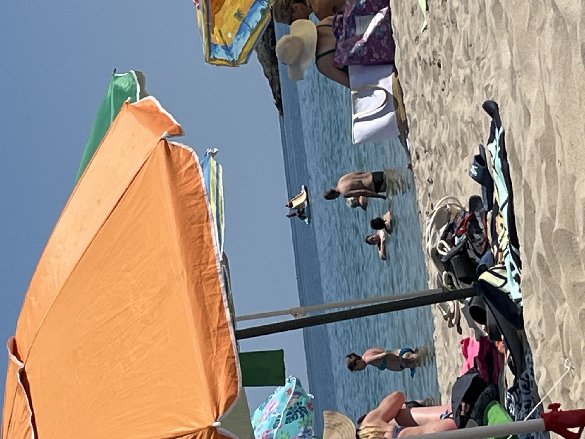 The image depicts a sunny day at a beach in Kiten, Bulgaria. In the foreground, a beach umbrella and various beach items, including towels, bags, and footwear, are scattered on the sand. Several people are visible in the shallow water, some swimming and others standing. In the background, a small water slide with two people on it is visible in the sea, and a boat is further out. The sea is calm and the sky is clear blue.