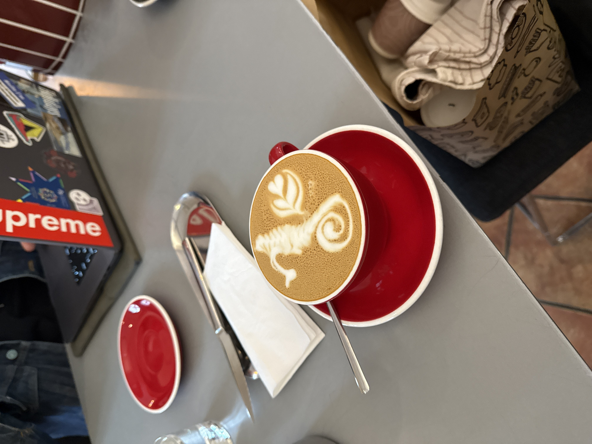 The image displays an overhead, slightly angled view of a grey tabletop within a cafe setting. The central subject is a red ceramic coffee cup with a white interior rim, containing latte art that forms a detailed seahorse figure on the left and two stylized heart-shaped leaf patterns on the right. This cup rests on a matching red saucer with a white rim. A silver spoon with a long handle is positioned within the cup, extending towards the viewer.
To the upper left of the coffee cup, an empty small red ceramic plate with a white rim is present. Further left, a clear glass, partially filled with liquid, is visible. To the left of the coffee cup, a folded white paper napkin lies underneath a silver butter knife. In the far upper left portion of the image, the shoulder and arm of an individual wearing a denim jacket are visible. Directly behind the red plate, an open laptop, identified as a MacBook, rests on a dark green or grey mat. The laptop's screen bezel features a prominent vertical red "Supreme" sticker, and its lid is adorned with various other smaller stickers.
In the lower right foreground, positioned next to the table, a brown paper bag with black line art depicting various kitchenware and food items is observed. Inside this bag, rolled fabric items, including a striped cloth or towel, are visible alongside other indistinct objects. The floor beneath the table is tiled with terracotta-colored squares. The specific location within Belgrade, Serbia, is not discernible from the visual information in the image.