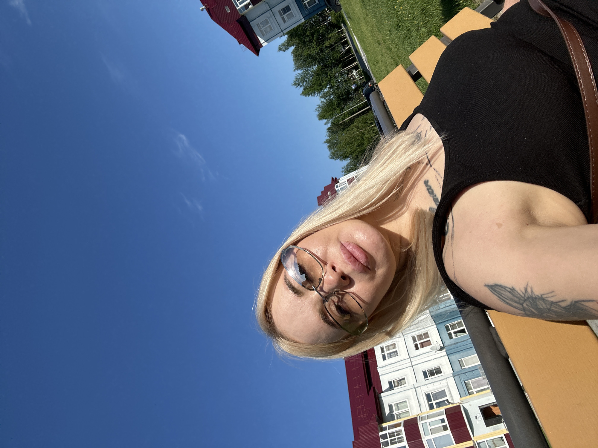 The image shows a woman with blonde hair and glasses, wearing a black top, sitting on a park bench outdoors. She is looking at the camera, and her expression is neutral. The background features a clear blue sky, green grass, trees, and buildings that appear to be residential apartment buildings with red and blue facades. The sun is shining brightly, suggesting it is daytime, likely late morning or afternoon. The woman has tattoos on her arm. There is no visible text in the image. The scene appears to be a casual outdoor setting on a sunny day.