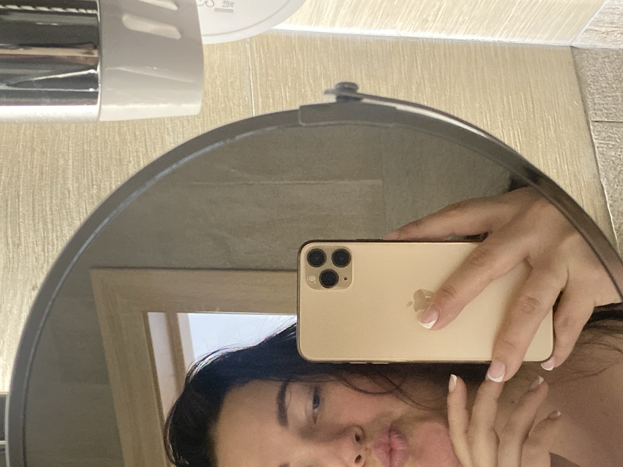A woman takes a selfie in a circular mirror. She is holding a gold iPhone with a prominent Apple logo on the back. Her face and hands are visible in the reflection, and she has French-manicured nails. The environment appears to be a bathroom, with a cylindrical object, possibly a soap dispenser, mounted on the wall above the mirror. The background reveals tiled walls and a partially visible wooden frame reflected in the mirror. There is no information to support the location being Sochi, Russia.