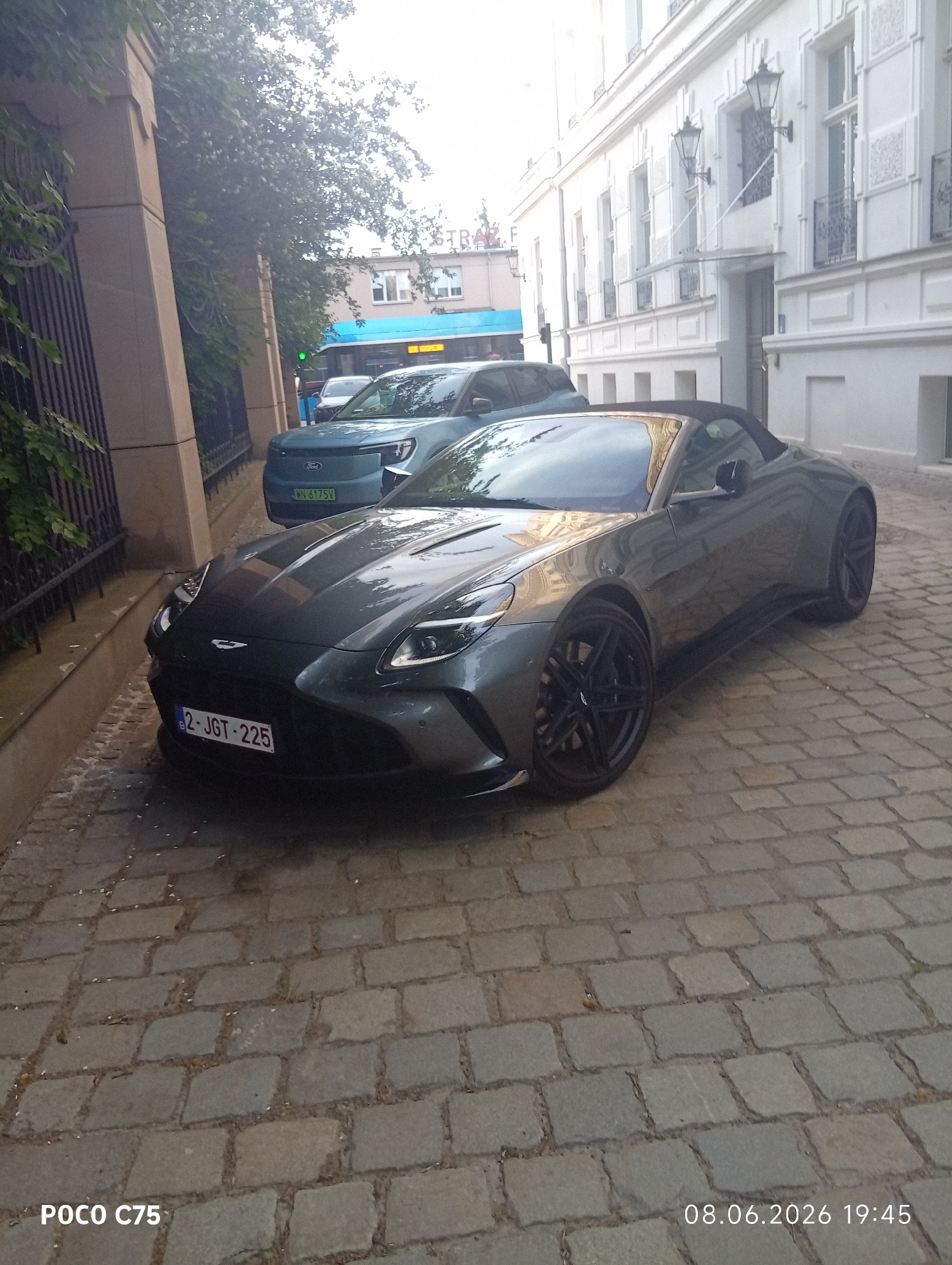 In Wrocław, Poland, a dark grey convertible Aston Martin Vantage is parked on a cobblestone street. The vehicle's license plate reads "2 JGT 225". Behind the Aston Martin, a teal Ford Explorer is visible, its license plate displaying "WN 6175V". The background includes a building with white facade and windows, and some trees and greenery on the left. A bus is also partially visible. The scene is captured from a low angle, with the timestamp "08.06.2026 19:45" in the lower right corner, and "POCO C75" in the lower left.