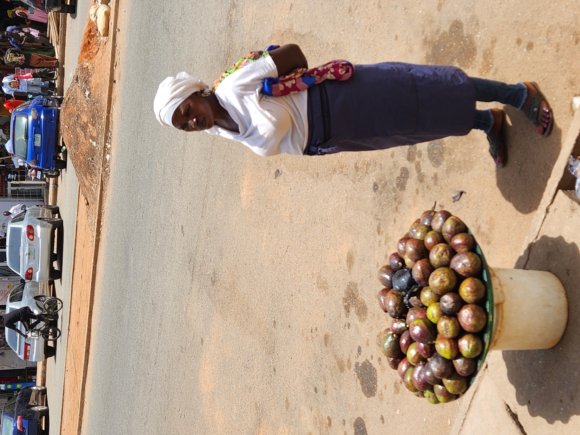 On a sunny day, in the city of Unknown, Unknown, a woman stands beside a bucket of fruit on a roadside. She wears a white headscarf, a white t-shirt with a colorful cloth slung over her shoulder, a blue skirt, jeans, and sandals with painted toenails. The bucket is filled with small, round fruits, ranging in color from green to red to dark purple. The road is asphalt, and there are several cars parked along the side of the road in the background. A group of people is visible near the cars.