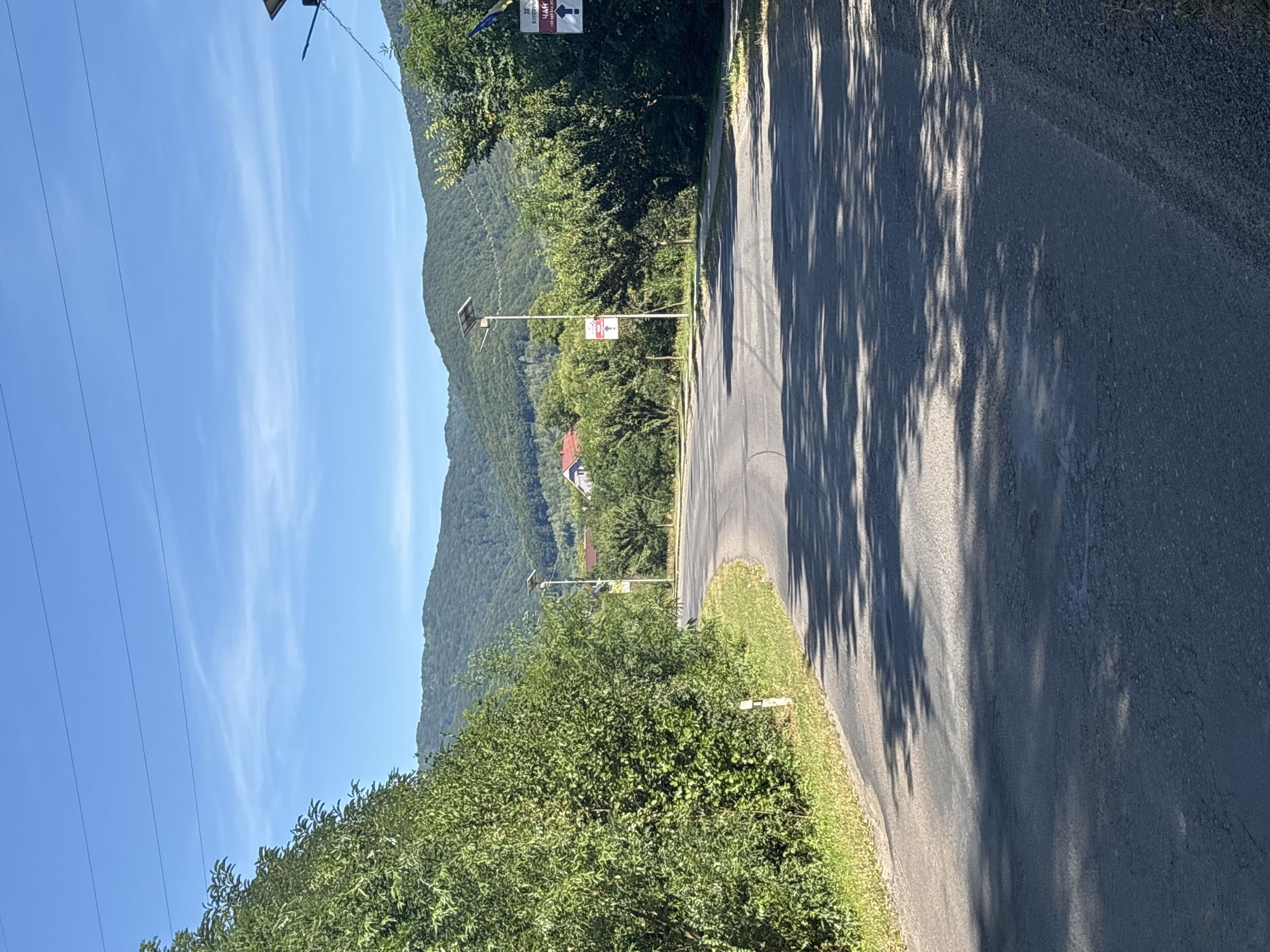 The image depicts a rural road in Сонячне, Ukraine, curving to the left and leading towards a distant village nestled among green, tree-covered hills under a bright blue sky with scattered white clouds. The road is paved with asphalt and shows signs of wear, with patches and cracks visible. Sunlight casts dappled shadows of trees onto the road surface.
On the left side of the road, dense green foliage from trees and bushes lines the shoulder, with a white marker post visible. On the right side, more trees form a dense barrier, and a few solar-powered streetlights are positioned along the road. Further down the road, in the village, a few houses with red roofs are visible. There are also signs along the road; one on the right prominently displays "КАІЕРЫНА" with a phone number and an arrow pointing to the left. Another smaller sign with an arrow is also visible further down on the right. The overall scene is one of a tranquil, sunny day in a mountainous rural area.