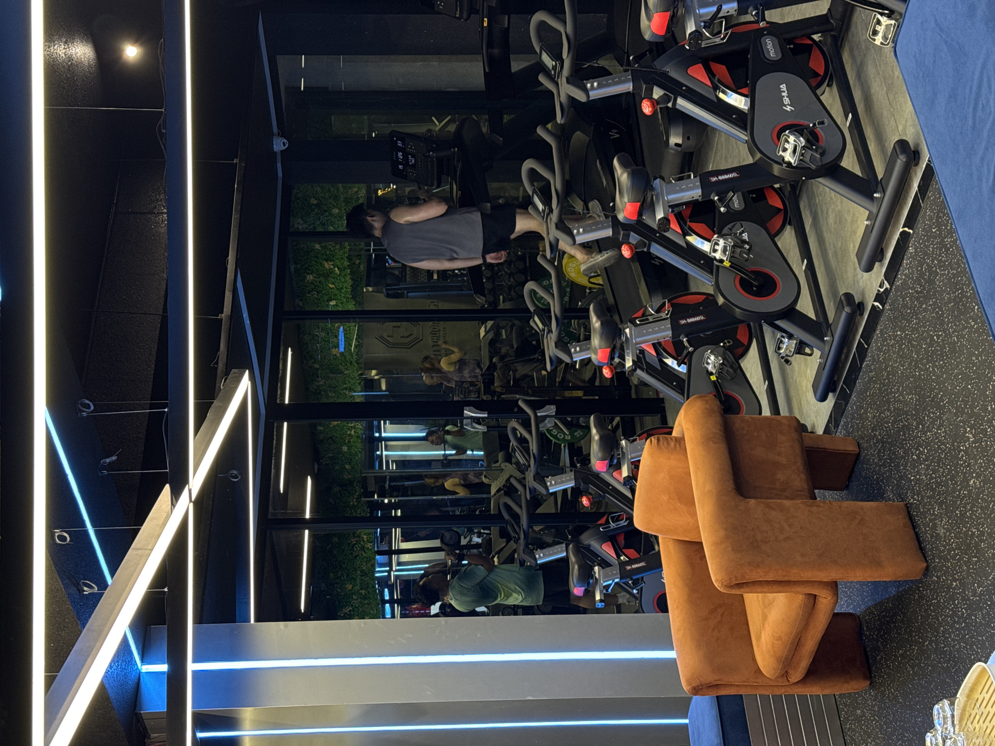 The image depicts the interior of a modern gym, likely located in Nam Nha Trang Ward, Vietnam. In the foreground, a striking, burnt-orange armchair with a unique, sculptural design sits on a black rubber gym floor. To the right, several stationary bicycles, likely for spinning classes, are arranged in a row. These bikes are black with red accents and feature digital displays.
In the mid-ground, through a glass partition, a man wearing a grey tank top and black shorts is jogging on a treadmill. His reflection is visible in the glass. Other gym equipment, including weights and machines, are faintly visible in the background through the mirrored walls.
The gym's ceiling features prominent, modern lighting, including long, linear white and blue LED strips. The overall ambiance is sleek and contemporary, suggesting a well-equipped fitness facility. The presence of multiple treadmills and stationary bikes indicates it's a popular spot for cardio workouts.
