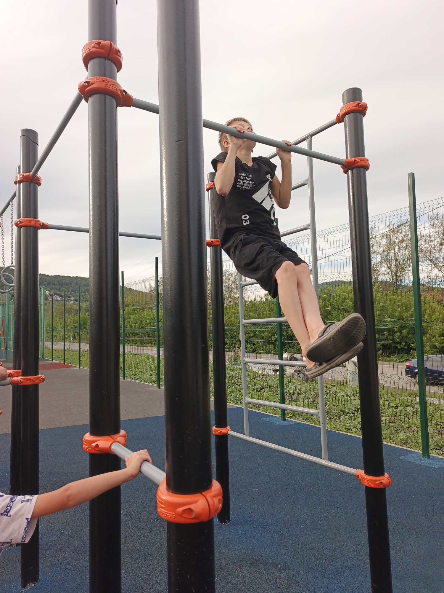 A young boy with short brown hair, wearing a black t-shirt and black shorts, is performing a pull-up on a horizontal bar at an outdoor calisthenics gym. His feet are slightly bent at the knees and his body is angled as he pulls himself up. Another child's arm, wearing a white t-shirt with a blue pattern, is visible on the left side of the frame, holding onto a horizontal bar. The calisthenics equipment consists of black metal poles with bright orange connectors and silver bars. The ground is a dark blue rubberized surface. In the background, a green metal fence encloses the area, beyond which is a road with a blue car and lush green trees and hills. The sky is overcast with a pale grey cloud cover. This scene is located at the children's playground in the city of Zlatoust, Russia, specifically at 67 Kalinina Street, near the park.