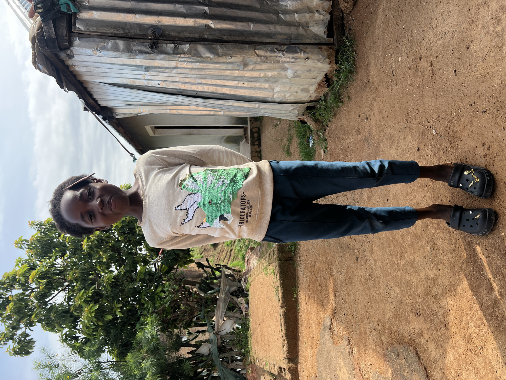 A young Black child stands in a dirt yard in front of a corrugated metal building, likely in Jos, Nigeria. The child is facing the camera and smiling slightly, wearing a light-colored long-sleeved shirt with a sequined green Triceratops graphic and dark blue pants. The child is also wearing black Croc-style shoes adorned with small charms. To the child's left, a small, raised, brick-like structure is visible, covered in dirt and sparse grass. Behind the child and to the left, lush green trees and foliage are present. The building behind the child appears to be a simple structure with a metal roof and corrugated metal siding, possibly a dwelling or storage area. The ground is a mix of packed dirt and sand. The sky is overcast with some visible clouds. The overall scene depicts a rural or semi-urban environment.