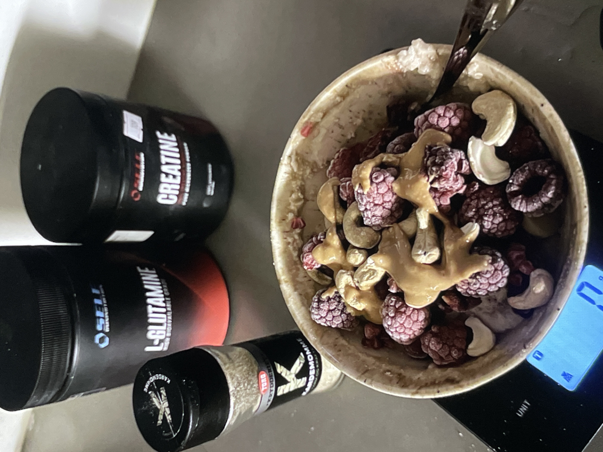 In Neubrandenburg, Germany, a close-up, top-down view captures a breakfast or post-workout meal preparation scene. Dominating the frame is a rustic, speckled bowl filled with frozen raspberries, whole cashews, and a drizzle of peanut butter. To the left of the bowl, a glass spice grinder with a black lid contains what appears to be cardamom, labeled "KARDEMOMME" and "TORG KRYDDERIER". In the background, two large black tubs of nutritional supplements stand prominently. The left tub, predominantly black with red and white accents, is labeled "L-GLUTAMINE HIGH BIOAVAILABLE CORE". The right tub, also black, is clearly marked with "CREATINE PURE MICRONIZED POWDER". The meal is positioned on a black digital scale, displaying a weight of "17" grams. The overall environment appears to be a kitchen counter.