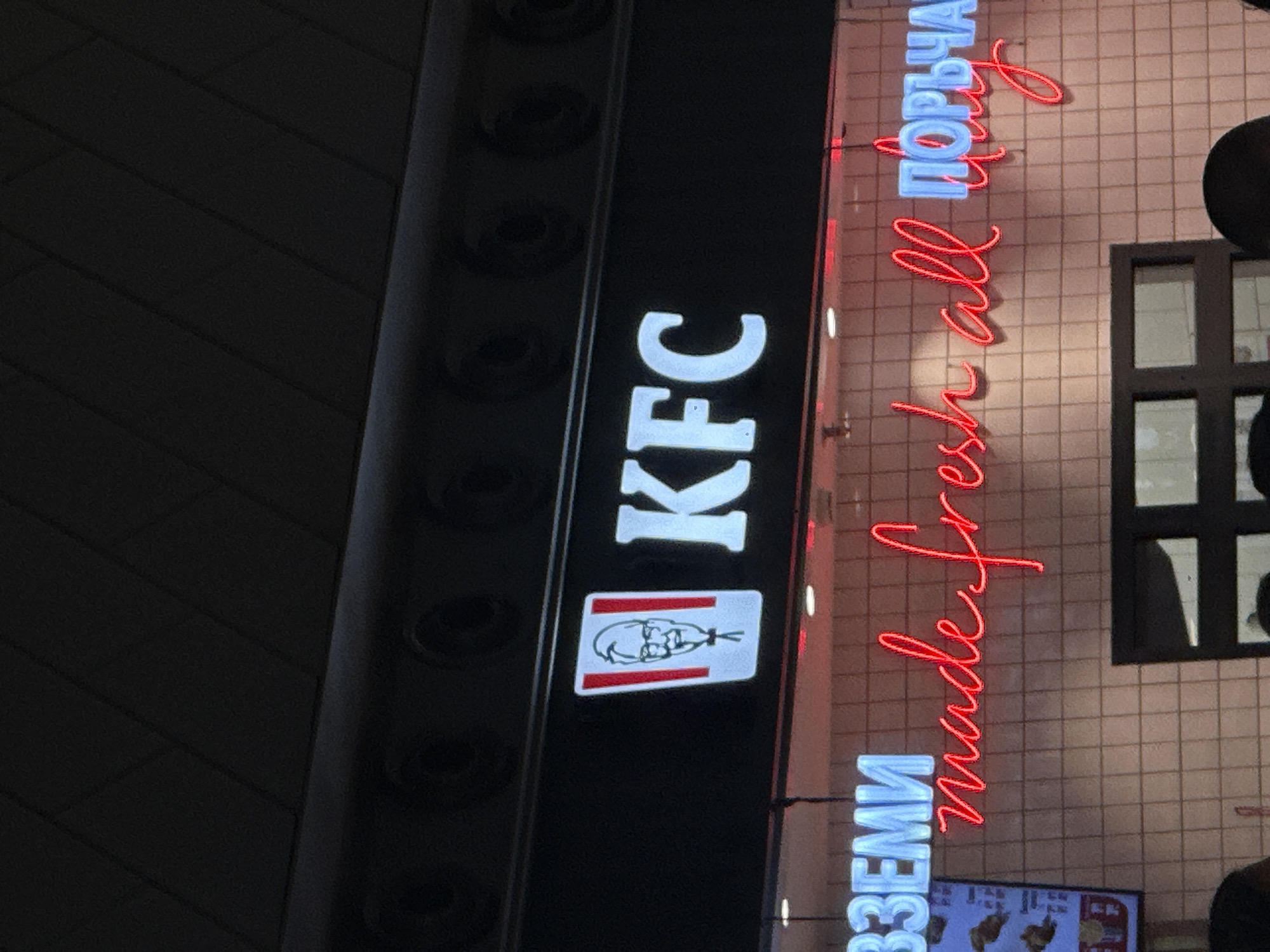 The image shows a nighttime view of a KFC restaurant sign in Sofia, Bulgaria. The sign is vertically oriented, displaying the KFC logo and text "KFC" in white against a black background. To the right of the sign is neon signage in red and light blue. In red, there's the text "made fresh all" written in a cursive font. Above this, in light blue is text written in Bulgarian Cyrillic script. The background consists of a tiled wall with a window.