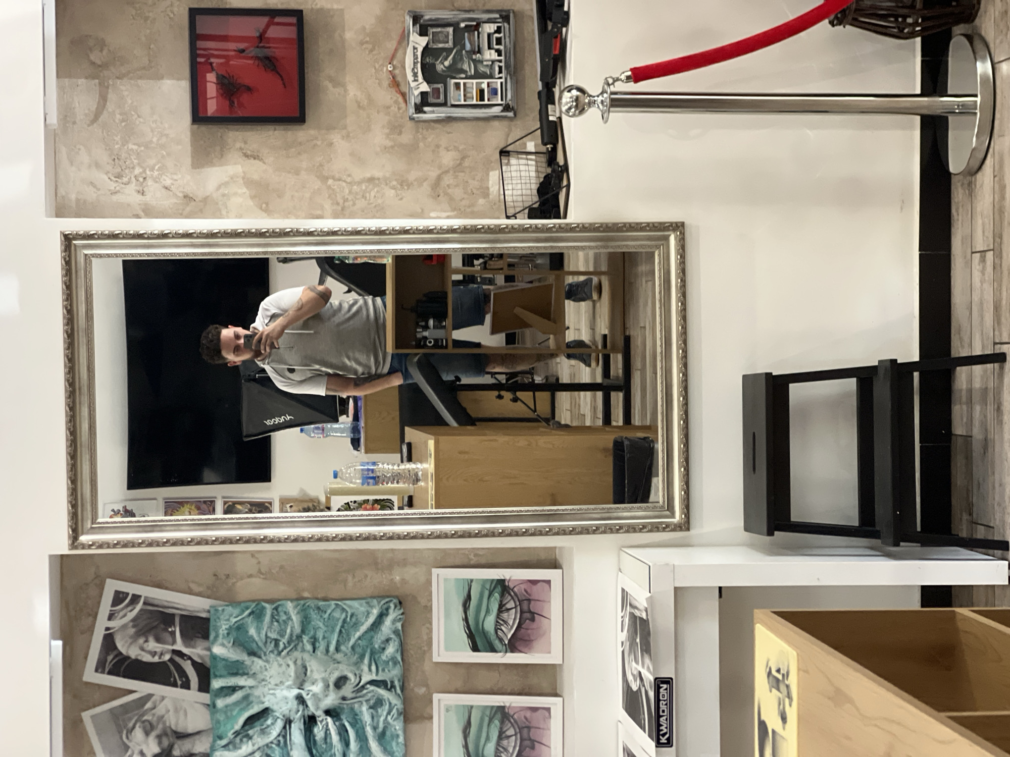 The image depicts an interior space, likely a commercial or artistic establishment, given the mix of art pieces and functional objects. A large mirror dominates the center, reflecting a man taking a picture with his phone, suggesting he is capturing the scene. The mirror's reflection shows a workspace with a desk, computer monitor, and lighting equipment. Underneath the mirror are a series of artworks, including black-and-white sketches, a blue sculpted piece, and eye drawings. To the right of the mirror is a stanchion with a red rope, a black stool, and a white shelving unit with light wooden boxes. The walls are textured, with decorative art pieces, including a framed red artwork and a shadow box containing various objects. The presence of the man, the artwork, and the functional items suggest the scene might be located in an art studio or a shop selling artistic creations. Given the level of detail in the image, it is difficult to determine if it is in the city of Sofia, Bulgaria, without additional contextual information.
