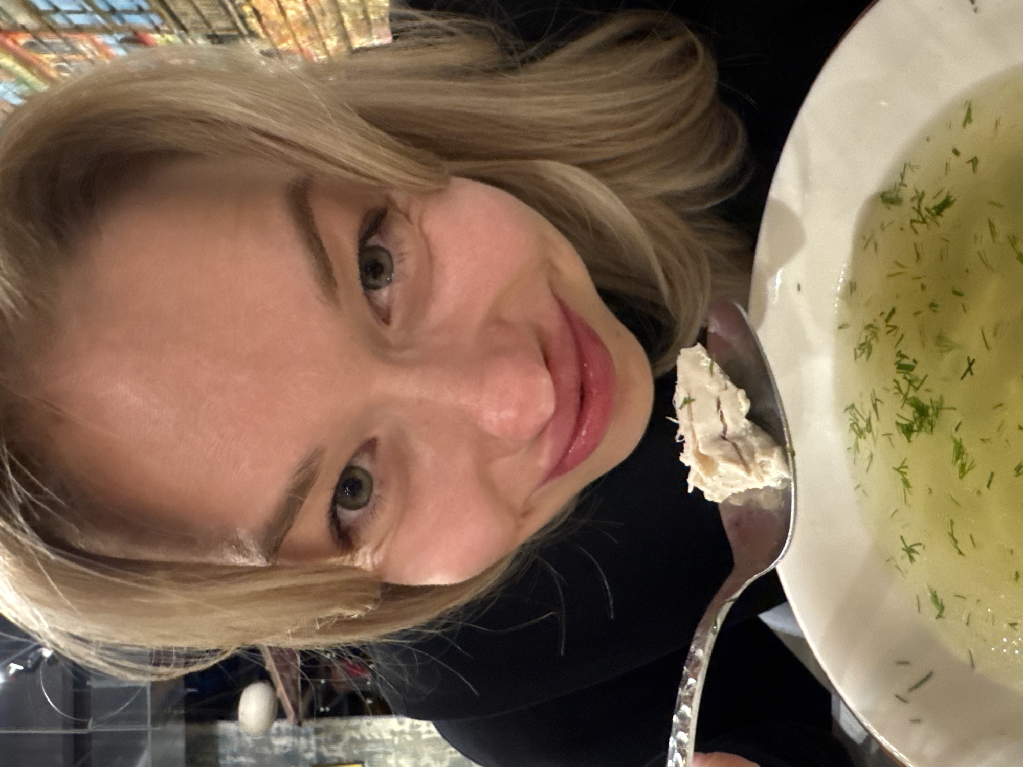 A close-up, high-angle image depicts an adult female with blonde hair and light-colored eyes smiling at the viewer while consuming soup in an indoor setting in Moscow, Russia. The woman, wearing a dark top, holds an ornate silver spoon containing a piece of light-colored meat over a white bowl filled with a clear, dill-garnished broth. Her lips are slightly parted. The background on the left is dark, featuring metallic elements and a light oval object. On the right, a vibrant, textured mural or wallpaper depicting a multi-colored building facade with windows provides an urban backdrop.