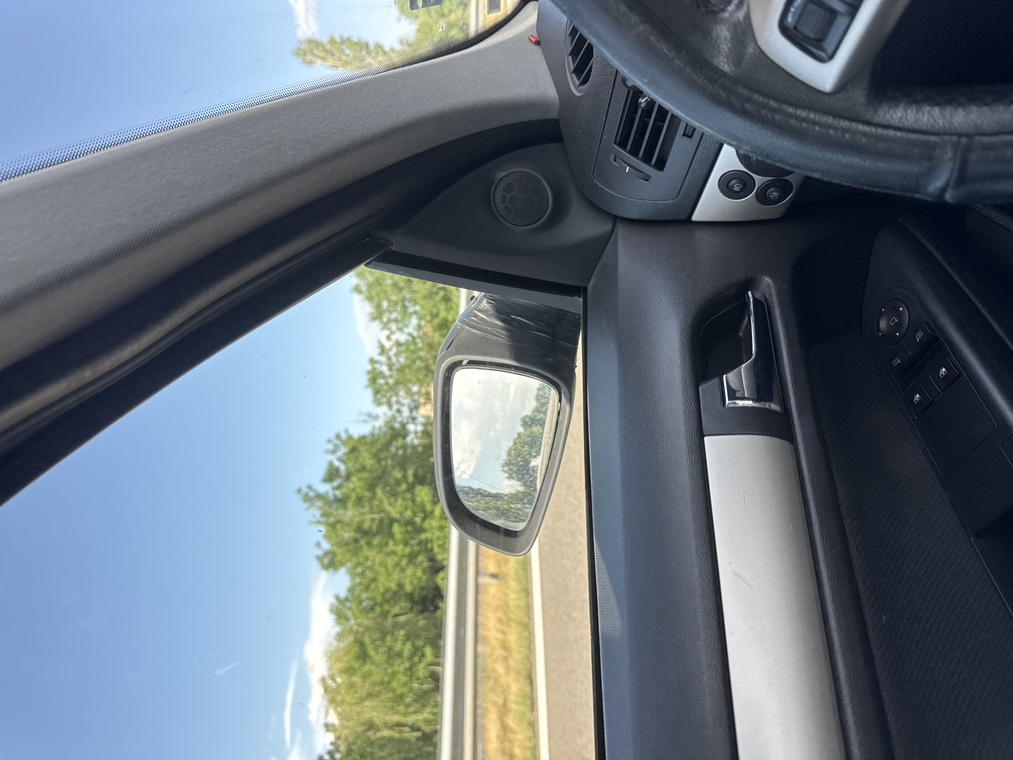 The image shows the interior of a car, specifically the driver's side. Visible elements include the steering wheel, air vents, door panel with controls, the side mirror reflecting a road and green trees, and the car window showing the sky. The location, Kalugerovo, Bulgaria, cannot be confirmed from the visual information in the image.