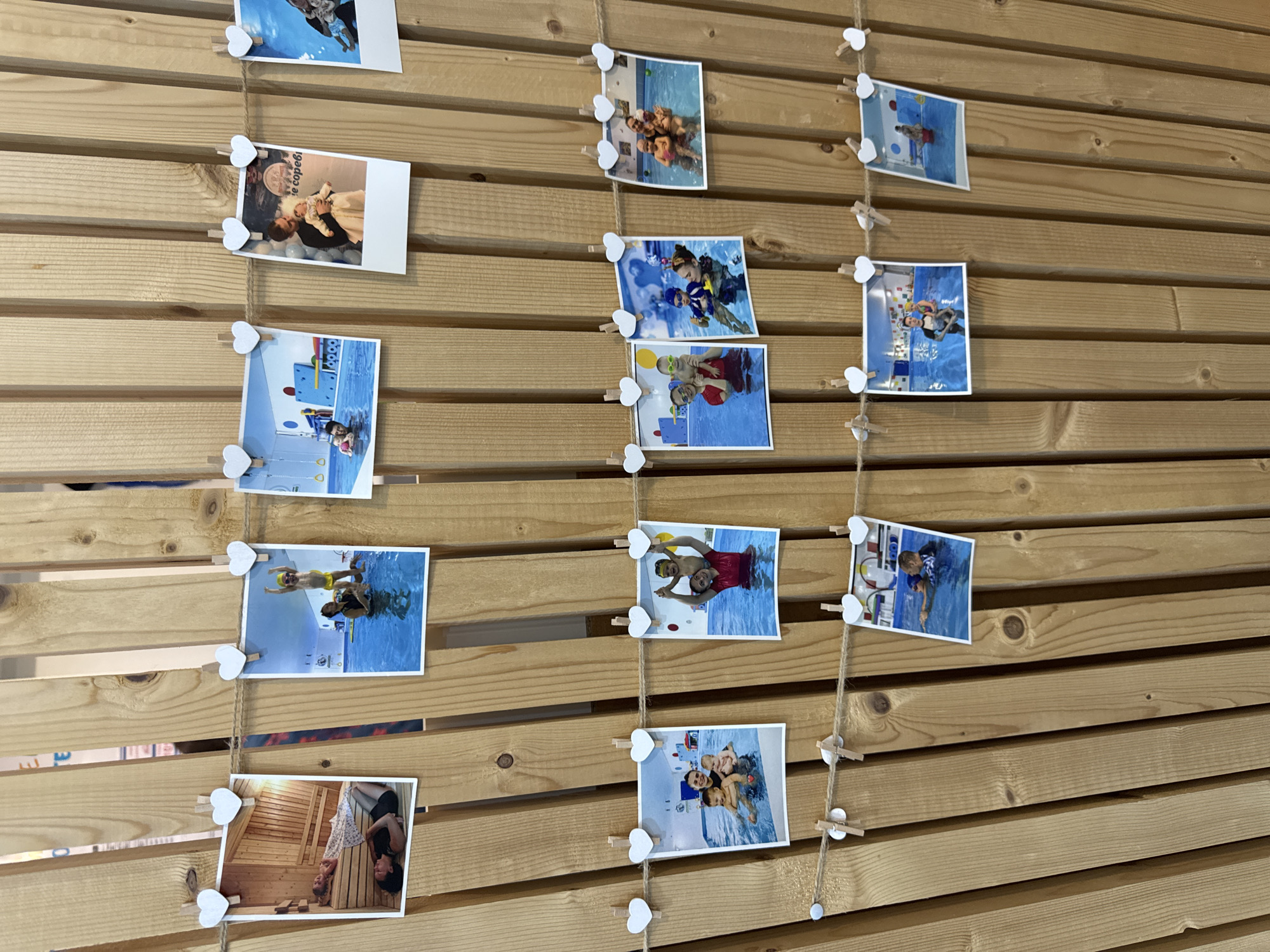 The image captures a display of personal photographs strung across a light-colored wooden slatted wall. Several strings of twine run vertically down the wall, each holding multiple instant-print style photographs with white borders.
The primary subjects in the photographs are children and adults, predominantly engaged in water-related activities. Many pictures show children in swimming pools, some appearing to be taking swimming lessons, others playing, or being held by adults. For example, one photo shows a child wearing a yellow swim cap and trunks in the water, while another depicts an adult holding two young children in a pool.
Beyond the pool scenes, one photograph features an adult holding a baby amidst balloons, suggesting a celebration or special occasion. Another distinct image shows an adult and a child inside what appears to be a wooden sauna or steam room.
The photographs are attached to the twine using small, decorative wooden clothespins, many of which are shaped like white hearts, adding a sentimental touch to the display. The wooden wall itself consists of horizontal slats, creating a warm and natural backdrop for the memories.
On one of the photos, the Russian phrase "В бассейне!" (meaning "In the pool!") is visible, printed upside down on the image. The overall arrangement suggests a collection of happy memories, likely from a family or children's recreational center, swimming school, or a similar facility, given the context of Sochi, Russia.