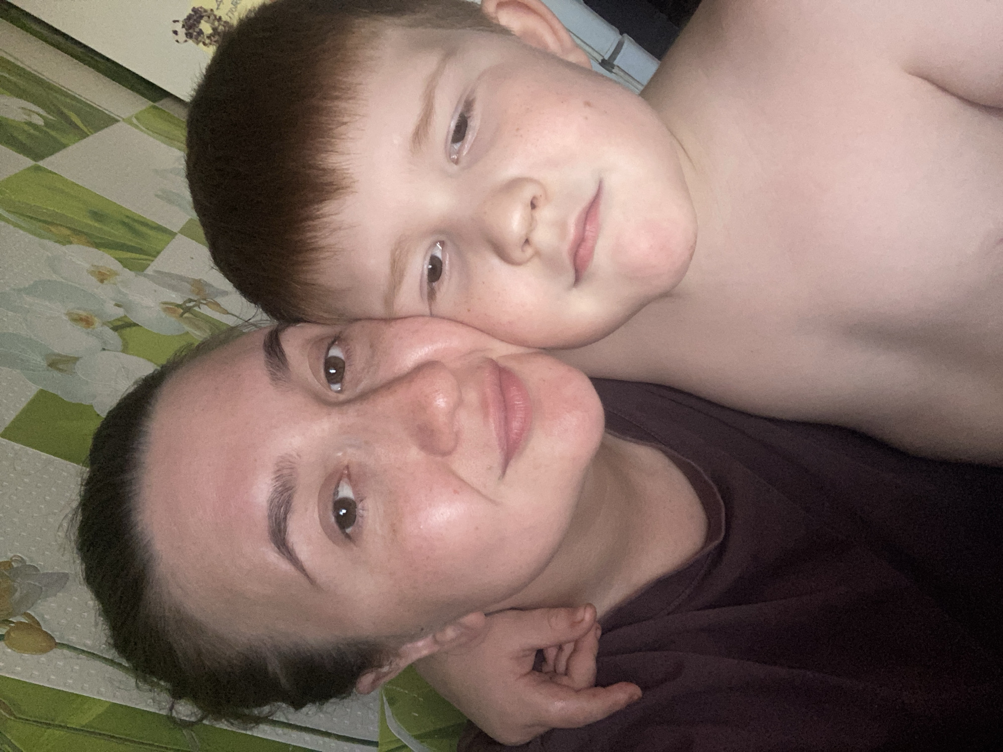 A close-up, frontal, eye-level selfie shows a woman and a young boy. The woman, on the left, has brown hair pulled back, brown eyes, and fair skin with freckles. She is wearing a dark maroon t-shirt. Her left arm is around the boy, and her hand is visible on his neck. The boy, on the right, has reddish-brown hair and fair skin with freckles. He is shirtless and his left arm is resting on the woman's shoulder. They are positioned in front of a wall with a floral wallpaper pattern. The image is brightly lit, likely from an artificial source. The overall impression is one of intimacy and affection. The location is described as the city of Chaikovsky, Russia.