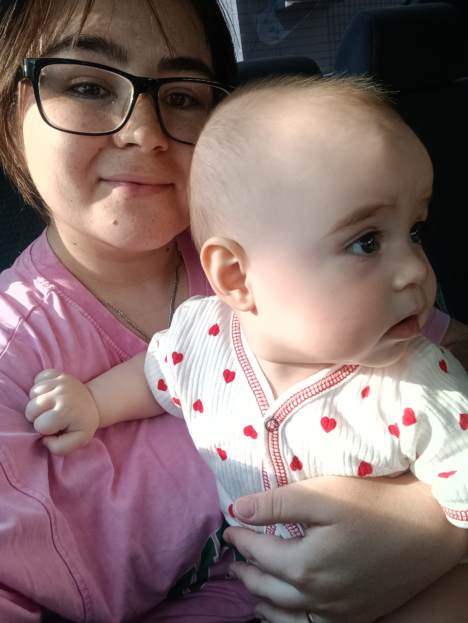 A medium shot captures a woman and a baby, likely inside a vehicle, as inferred from the seat visible in the background. The woman, wearing glasses and a pink t-shirt, is positioned slightly behind the baby. The baby, dressed in a white onesie adorned with red hearts, is held by the woman. The baby is looking towards the right side of the frame. The location is not determinable from this image, and the city of Noviy, Russia, cannot be confirmed.