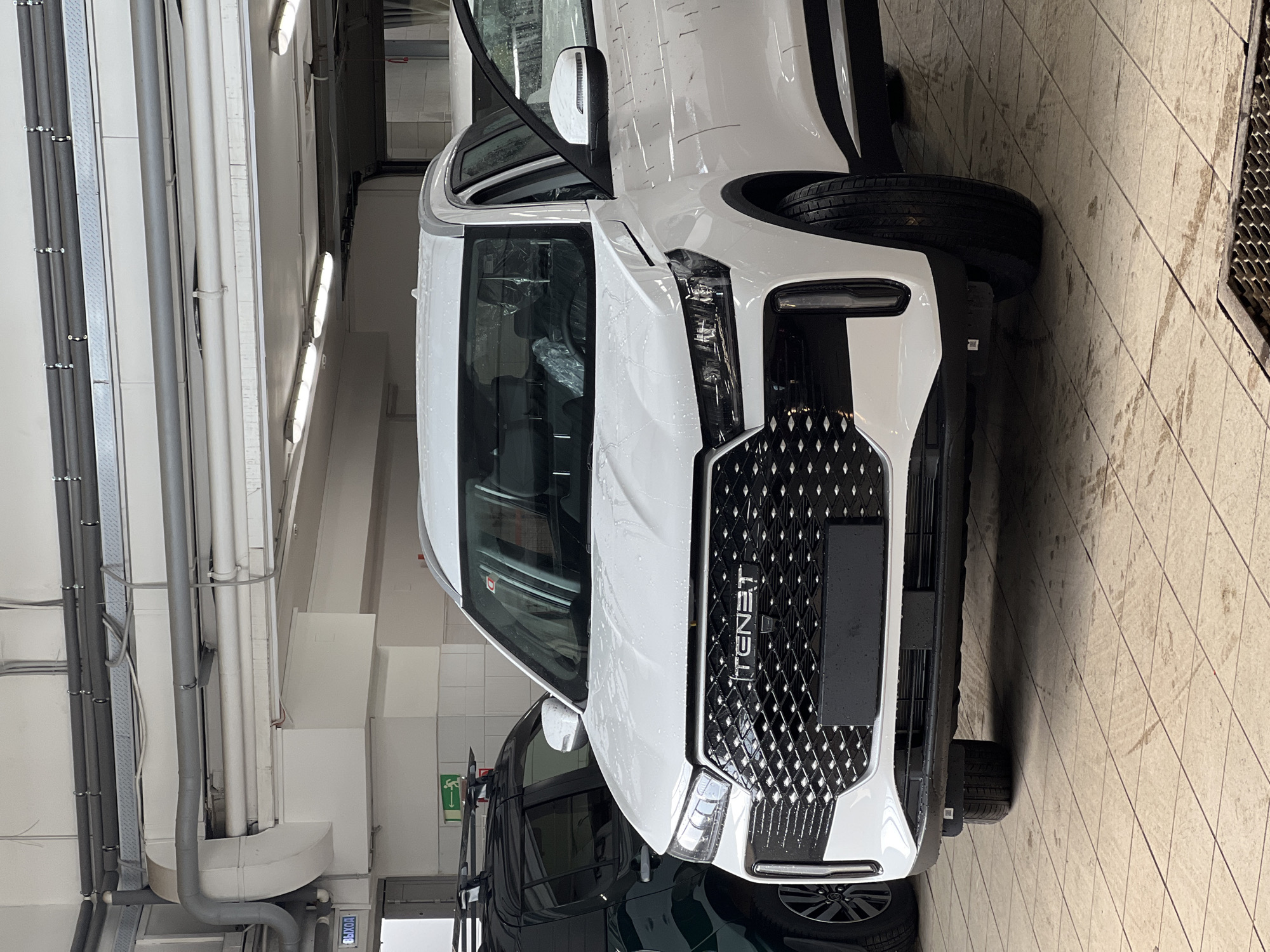 The image depicts the interior of a commercial or industrial facility, likely a car service center or showroom, situated in Izhevsk, Russia. A white SUV, identified by the "TENET" emblem on its prominent front grille, occupies the central and right foreground. The vehicle's surface, particularly the hood and roof, exhibits numerous water droplets, indicating recent exposure to water. The driver's side front door is open, revealing the interior where seats are covered in clear protective plastic. The front license plate area is a black, blank rectangle.
To the left and rear of the white SUV, a dark green or black SUV with a roof rack is partially visible.
The floor is covered with light-toned square tiles, showing signs of dampness in various areas. A metal grate, indicative of a drainage system, is present in the lower right foreground. The walls feature white rectangular tiling up to a certain height, transitioning to plain white painted surfaces above. Overhead, the ceiling structure reveals exposed utilities including multiple grey, insulated pipes, white rectangular air ducts, and rows of linear fluorescent light fixtures providing illumination.
Signage on the walls includes a blue illuminated sign with white Cyrillic text reading "ВЫХОД" (EXIT) on the upper left, and a green emergency exit pictogram sign (person running) on the wall behind the dark SUV. No individuals are visible within the scene.