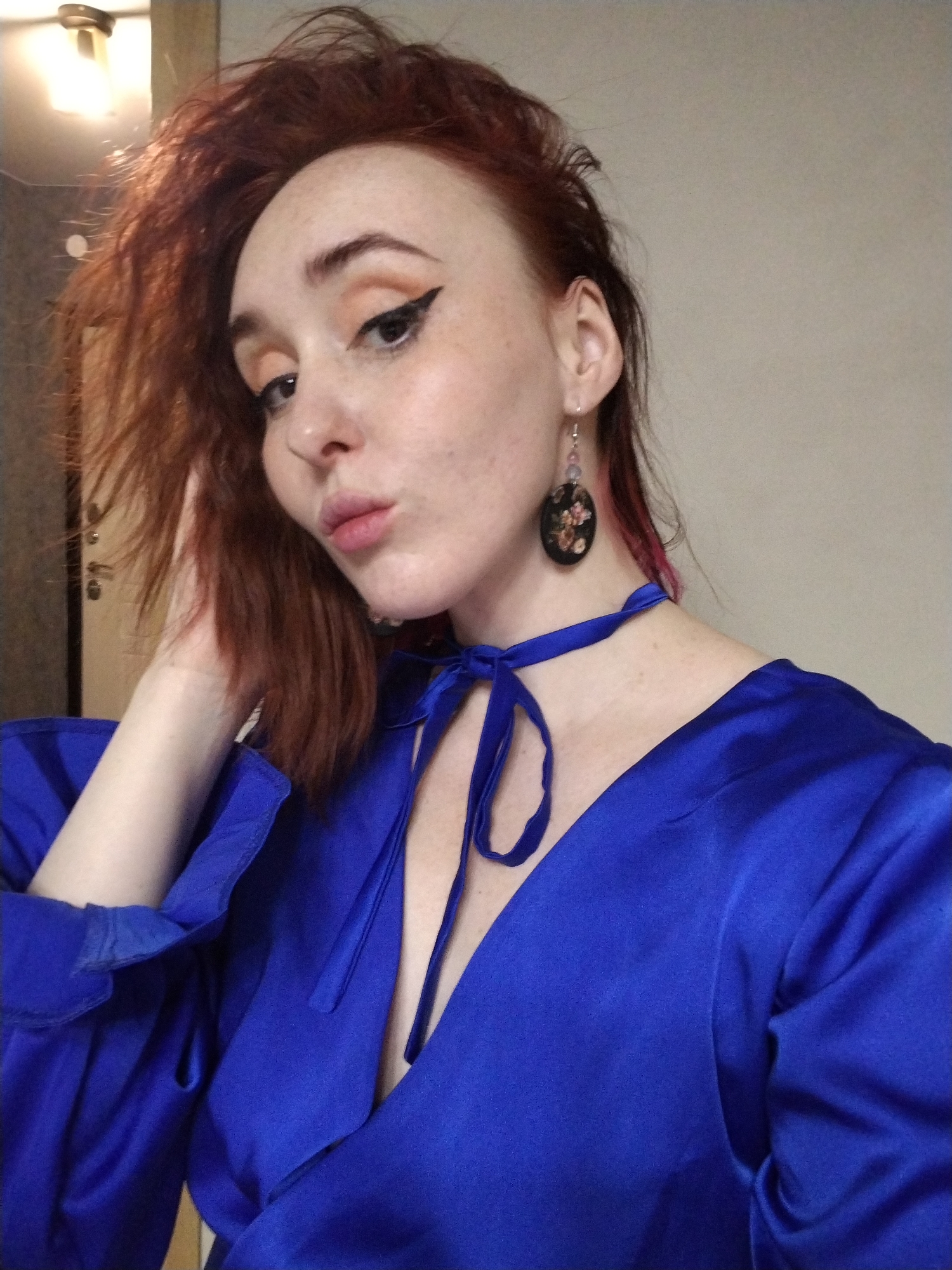 A fair-skinned woman with red hair and freckles is shown in a selfie, positioned slightly off-center. She is wearing a shiny blue wrap-style dress with a plunging neckline and puffy sleeves, and a matching blue ribbon choker. Her hair is somewhat messy, with some strands falling across her forehead. She is wearing eye makeup with a winged eyeliner. She has one earring with an oval pendant with a floral design. She is touching her hair with her left hand. The background is a neutral wall. The lighting is somewhat dim, with a light fixture visible in the top left corner of the frame. There is no indication of Izhevsk, Russia in the image.