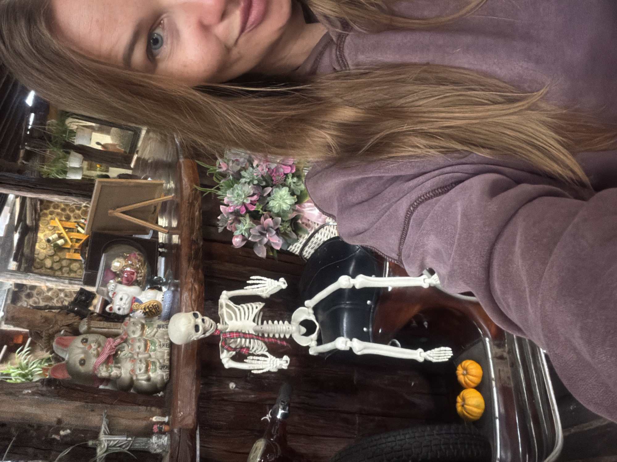 A young woman with blonde hair and blue eyes is taking a selfie in a dimly lit, rustic room. She is wearing a purple hoodie. To her left, a decorative skeleton is seated on what appears to be a motorcycle seat. The skeleton is wearing a red and black plaid scarf. In the background, there are several decorative cat figurines and plants. Two small orange pumpkins are placed on a metallic surface in the foreground. The overall ambiance suggests a quirky, possibly Halloween-themed, interior setting. The location is identified as Lâm Viên - Đà Lạt, Vietnam.
