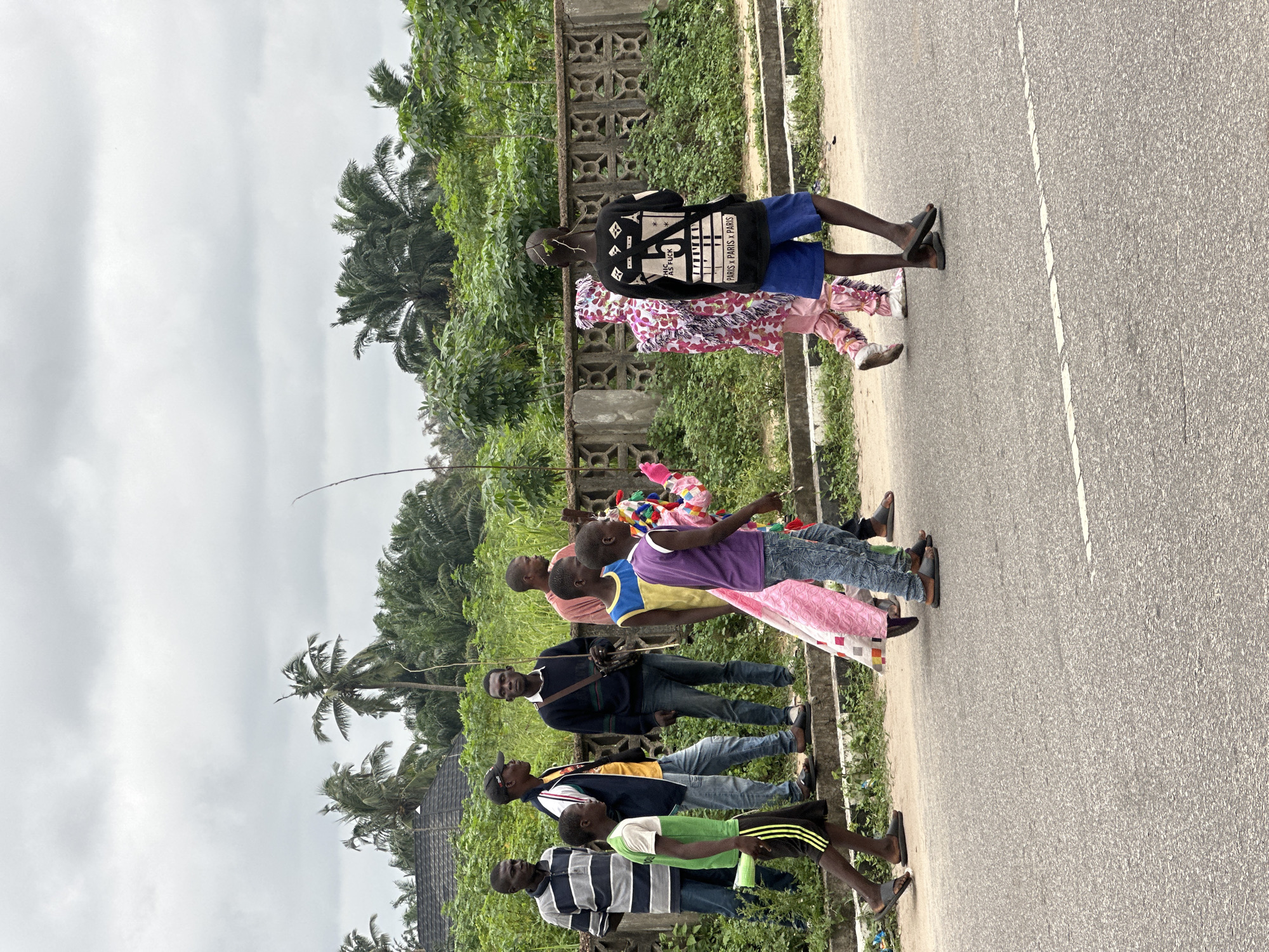 In Badagry, Nigeria, a group of people is walking along a paved road under an overcast sky. On the left, six men are standing and walking near a low, concrete wall overgrown with green foliage. The men are wearing casual clothing, including striped shirts, a t-shirt, a long-sleeved shirt, and a t-shirt with a jacket. One of the men is holding a stick. To the right of the men, three children are walking on the road. One child in the foreground is wearing a green t-shirt and black shorts. Behind him, a child in a purple t-shirt and jeans is walking, holding a colorful object. Further to the right, another child is wearing a pink and white costume and blue shorts. The background features palm trees and dense green vegetation behind the concrete wall. The road has a white dashed line marking the center.