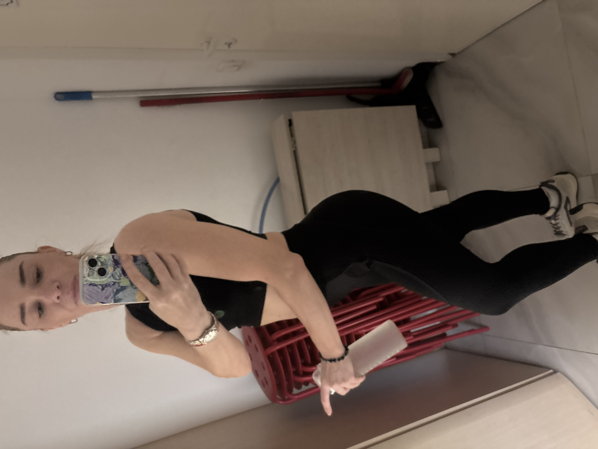 In an indoor, likely domestic or storage setting, a woman with blonde hair pulled back poses for a mirror selfie. She is wearing a black sports bra and black leggings, along with white and grey athletic shoes. On her left wrist, she wears a watch. In her left hand, she holds a clear water bottle. Behind her, a stack of red stools is visible. To her right, against a plain white wall, a mop with a blue head stands next to a wooden cabinet. The floor appears to be tiled. The lighting is artificial and somewhat dim. The background is cluttered with various objects, suggesting a functional space rather than a display area. The woman's expression is neutral as she looks into the mirror.