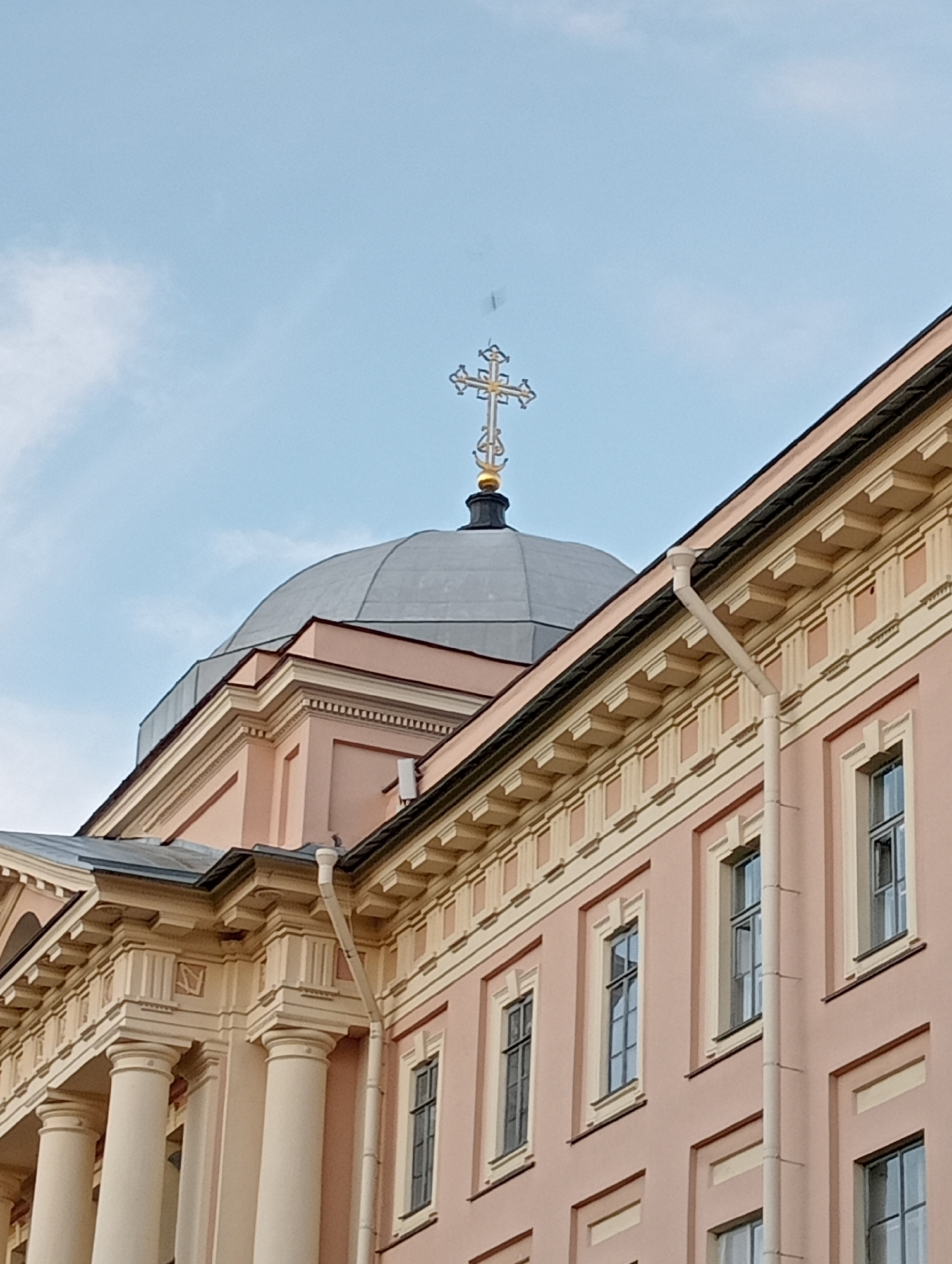 The image captures a detail of a pale pink building in Saint Petersburg, Russia, featuring a domed roof topped with a golden cross. The building exhibits classical architectural elements, including ornate cornices, pilasters, and columns supporting an upper section. Rain gutters are visible running down the side of the building. The sky above is a pale blue with a few wispy clouds. The perspective is from a low angle, looking upwards at the building's structure and its prominent religious symbol.