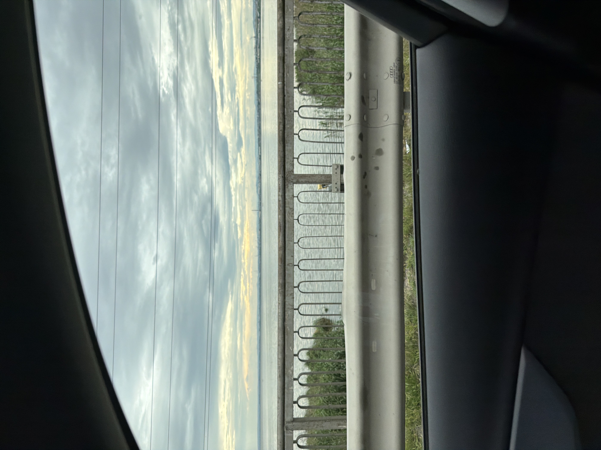 The image, likely taken from inside a vehicle, shows a partial view of a waterway. A gray metal guardrail runs parallel to the water. Beyond the rail, sparse vegetation grows along the water's edge. The water appears calm, reflecting the sky, which is mostly overcast with clouds. Power lines are visible above the water. The interior of the vehicle is partially visible in the foreground. The image has been taken in the city of Burgas, Bulgaria, based on the railing design and the landscape.