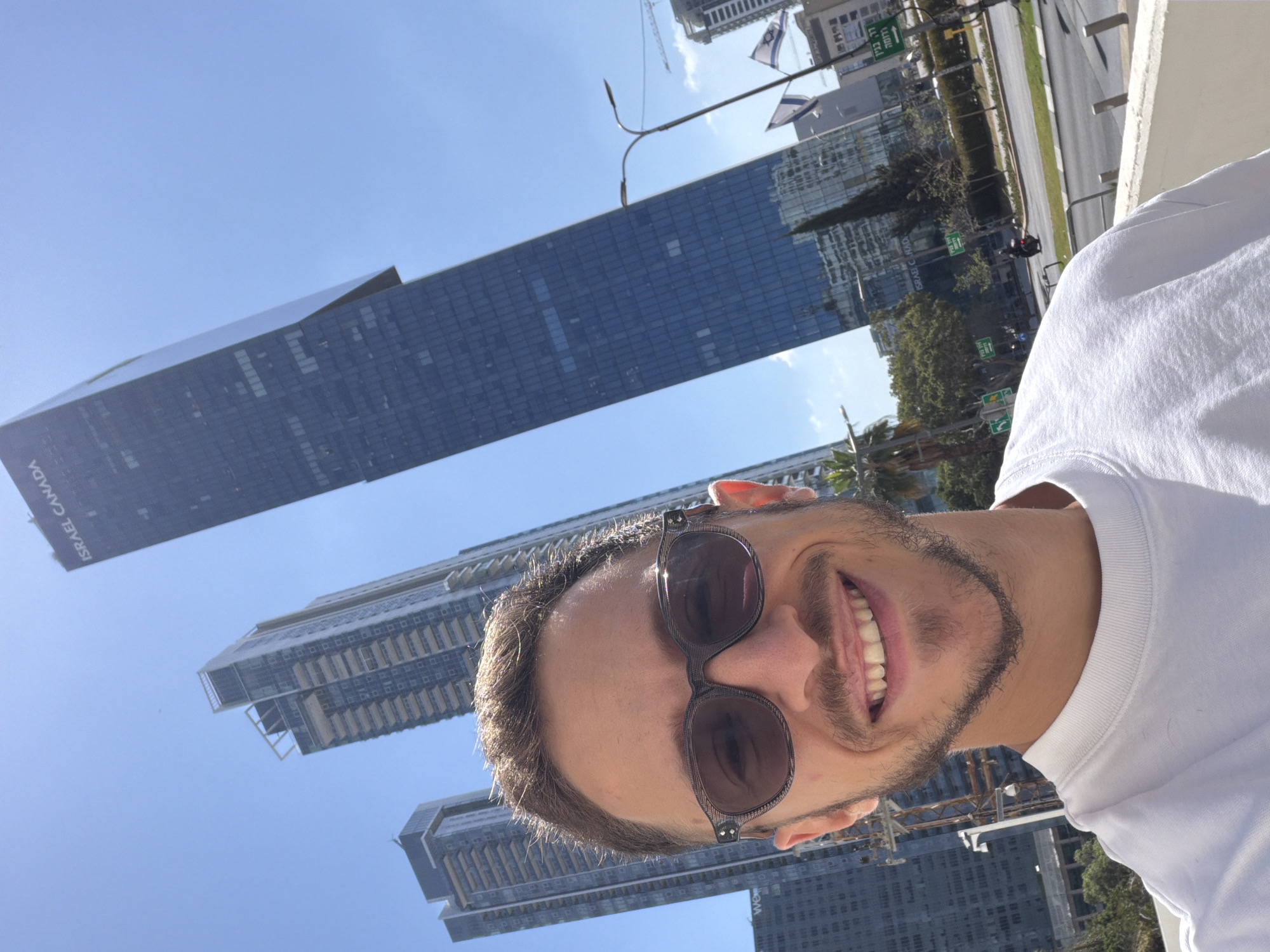 The image is a selfie taken from a low angle, showing a man wearing sunglasses with a white shirt and a beard with a smile. The background consists of modern skyscrapers, a clear blue sky and some construction cranes. The text "ISRAEL CANADA" is visible on one of the buildings. The location is likely Tel Aviv, Israel, as seen by the Israeli flags.