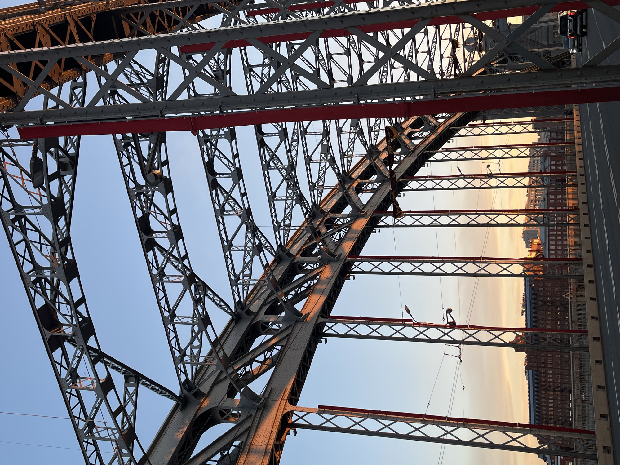 The image shows the truss structure of the Peter the Great Bridge in Saint Petersburg, Russia. The bridge is constructed of steel and features a complex network of interconnected beams and girders. The bridge appears to have a roadway alongside the metal structure. Red accents are painted along structural elements of the bridge. Beyond the bridge, buildings with windows are visible. The sky is light blue with a setting sun.