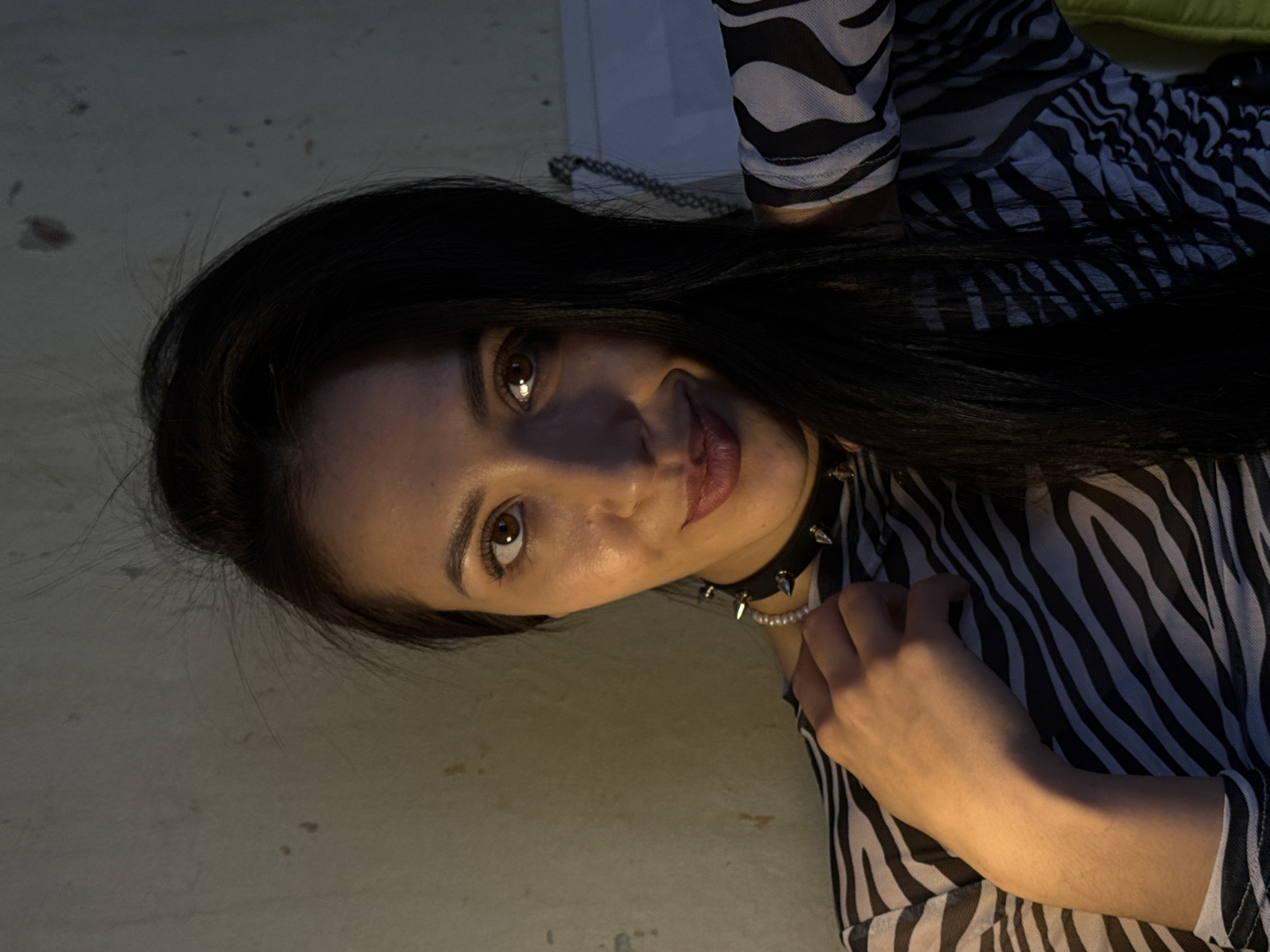 A woman with dark hair is positioned in the frame, angled slightly with her head to the left. She wears a zebra-striped top and a choker with spikes and pearls. The environment appears to be indoors, with a neutral-toned surface visible in the background. The information provided does not allow for the determination of a location in Votkinsk, Russia.
