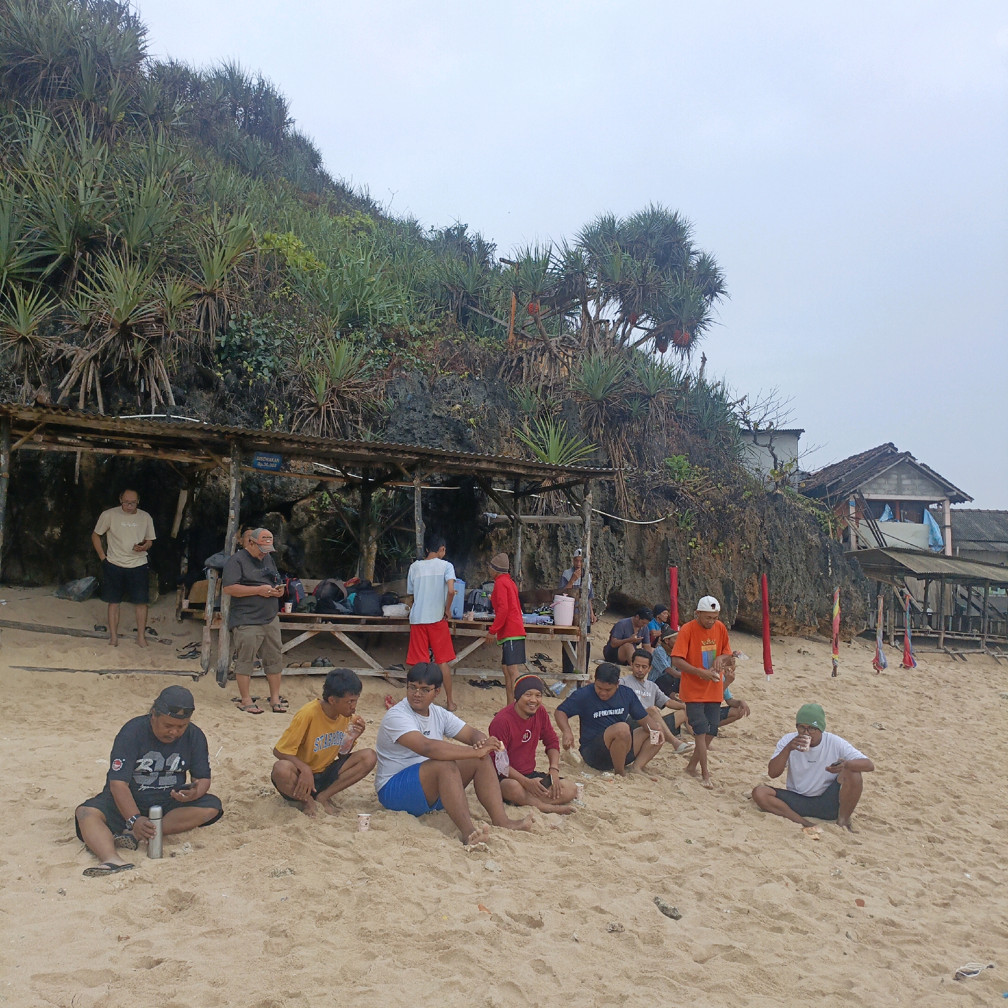 A group of approximately 15 men are gathered on a sandy beach near a rocky cliff face in Tepus, Indonesia. The scene appears to be under an overcast sky. Several men are seated on the sand, some in small groups, while others stand. In the background, a simple wooden structure with a corrugated metal roof serves as a shelter, with some individuals near or within it. A few bags and a cooler are visible near the shelter. The cliff face behind the shelter is covered in dense green vegetation. The men are dressed in casual clothing, including t-shirts, shorts, and hats. Some are holding drinks or food. The overall atmosphere suggests a relaxed outing or gathering at the beach.