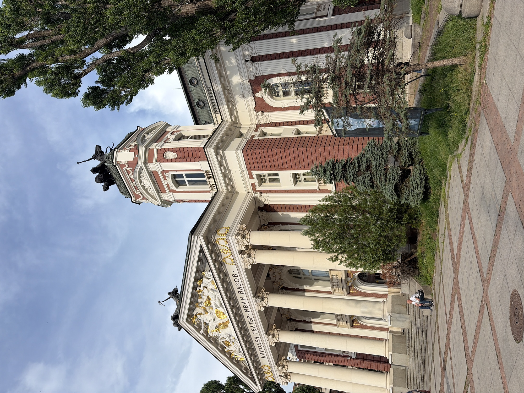 The Ivan Vazov National Theatre in Sofia, Bulgaria, is captured in this exterior shot on a partly cloudy day. The grand theatre, with its distinctive red brick facade and ornate white architectural elements, dominates the frame. A statue group is visible atop the central pediment, and a smaller sculptural element sits on a turret to the right. Several tall, mature trees with green foliage are present in the foreground and to the right of the building, casting some shadows. In front of the theatre, a paved area with a geometric pattern of square tiles and reddish brick borders leads up to a set of stairs. A man in a white t-shirt and dark pants is seated on a stone ledge to the left of the stairs, facing towards the theatre. Grass and some smaller evergreen trees are growing in patches along the base of the theatre and the edge of the paved area.