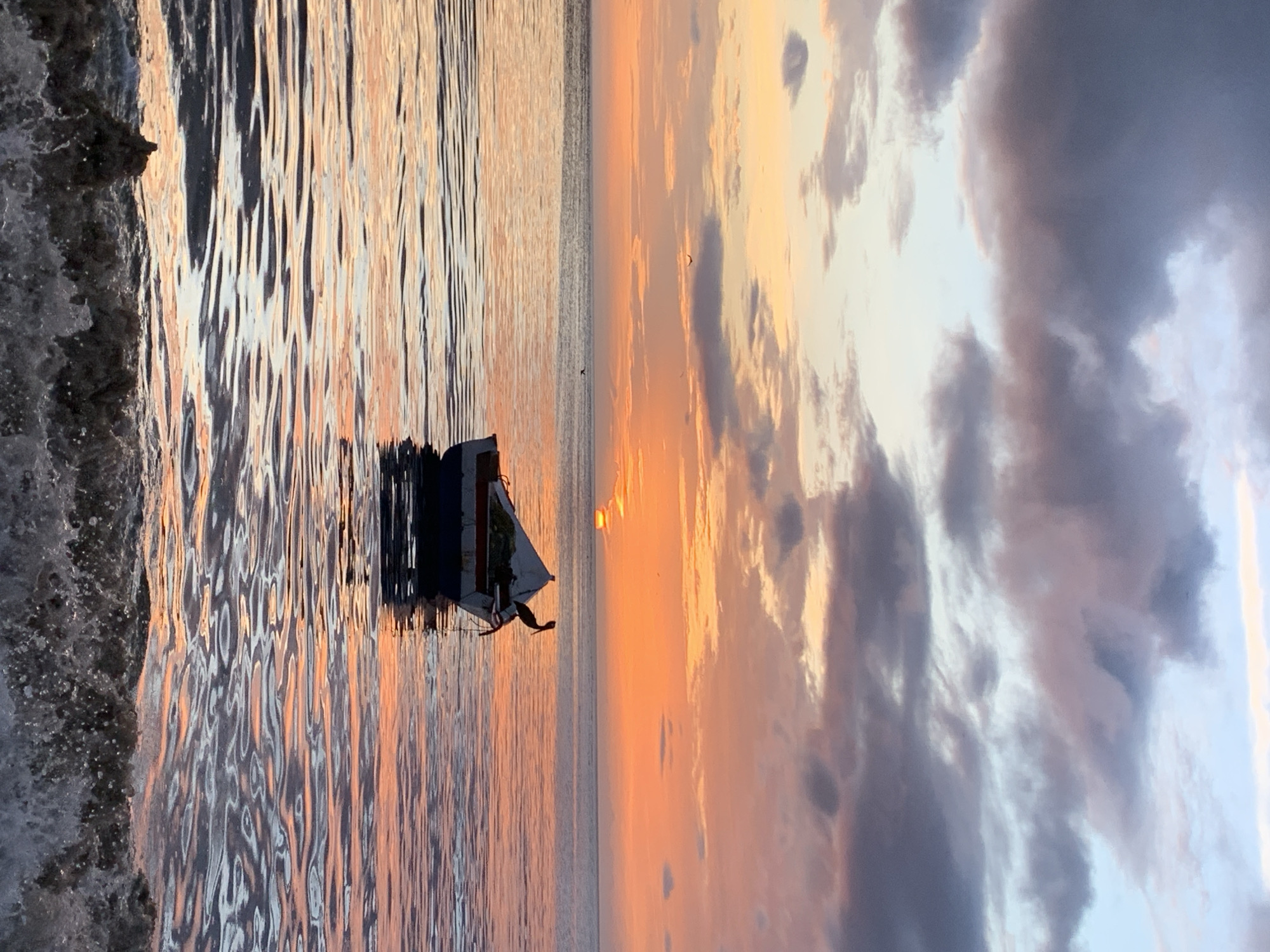This is a vertical, low-angle shot of a sunset over the ocean in Juan Griego, Venezuela. The sun is low on the horizon, casting a warm orange glow across the sky and reflecting on the rippling water. Dark, dramatic clouds fill the sky on the right side of the frame, with lighter clouds scattered throughout. In the foreground, waves crash against the shore on the left side of the image. A small boat, partially submerged, is visible in the water on the left side, with what appears to be a bird perched on it. The scene evokes a sense of peace and the beauty of nature. The time of day is sunset, and the weather appears partly cloudy with calm seas.