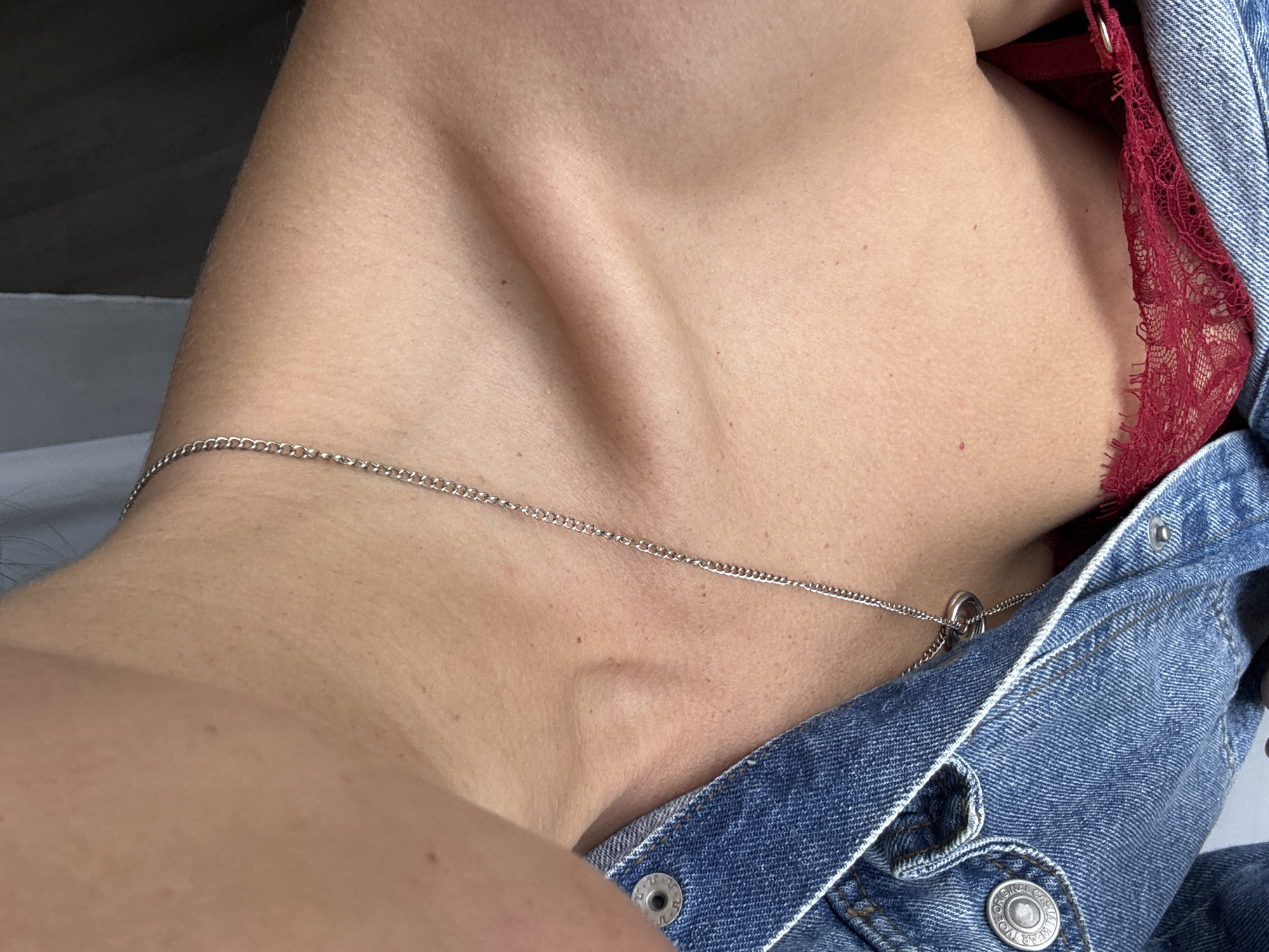 A close-up, high-angle shot focuses on the décolletage area of a person wearing a blue denim jacket and a red lace bra. A silver chain necklace with two circular pendants rests on the skin. The person's skin appears fair with a scattering of freckles and a few reddish moles. The background is blurred, with a dark gray and a light gray area visible. The image is cropped to exclude any facial features or full body.