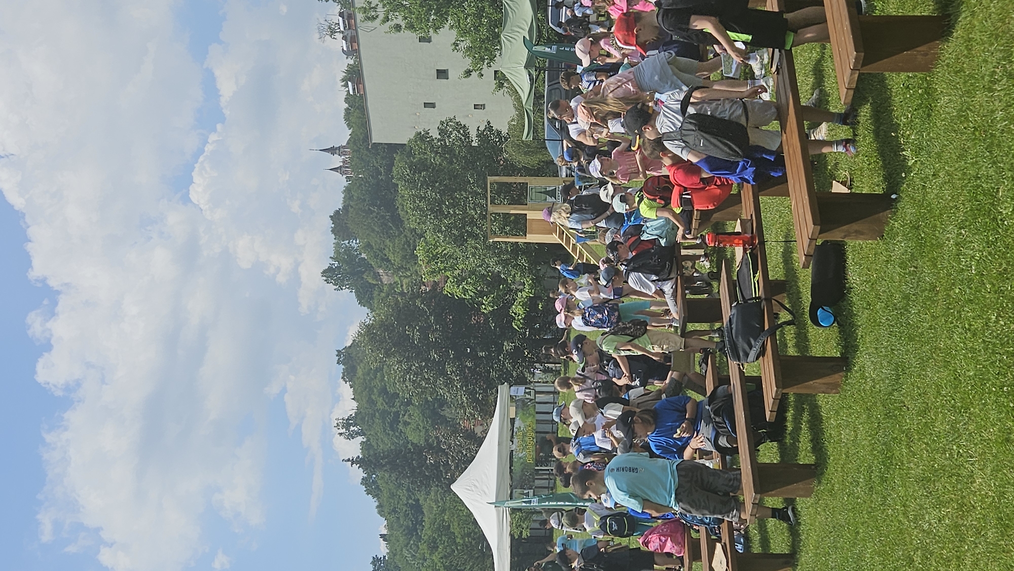 The image depicts an outdoor event, likely a festival or educational program, taking place on a sunny day in a grassy park. A large group of children, mostly wearing casual clothing and backpacks, are gathered around wooden benches and tents. Some children are sitting on the benches, while others are standing and interacting. A white tent with signage is visible on the left, and a green tent is present on the right. In the background, there are trees and buildings, including a church spire on a hilltop, suggesting an urban or suburban setting within the city of Prešov, Slovakia. The overall atmosphere appears to be lively and family-friendly, with many children participating in activities or observing.