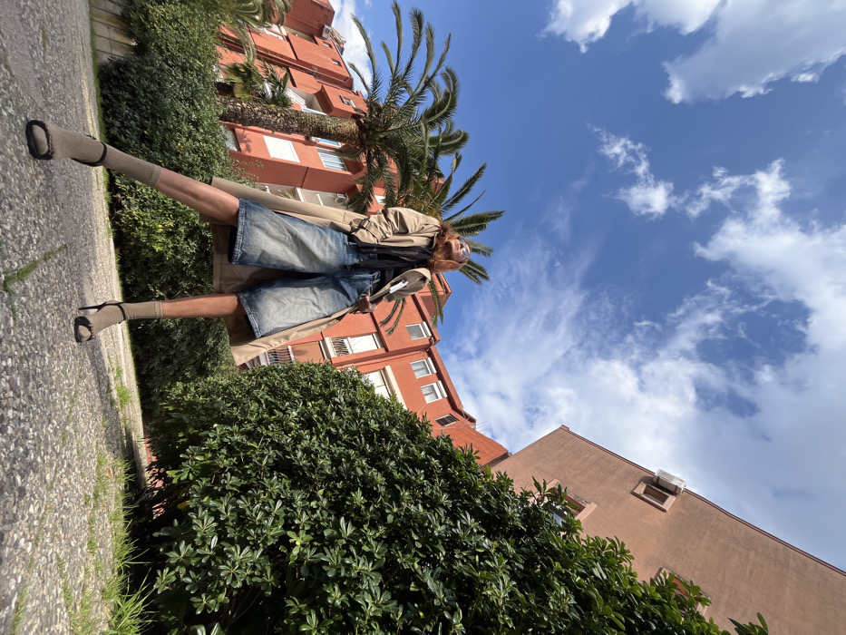 The image displays a female individual standing on a paved outdoor surface in the city of Budva, Montenegro, looking upward. She wears a beige trench coat over a dark top, light blue denim culottes, and black high-heeled sandals worn over light beige socks. The background features multi-story residential buildings with predominantly reddish-orange facades. To the left, a building shows multiple white-framed windows and balconies, with several tall palm trees visible behind it. To the right, another building of a slightly lighter reddish-orange hue has an exterior air conditioning unit mounted on an upper wall. Dense green hedges line the ground level, separating the paved area from the buildings. The ground is a light-colored aggregate pavement with minimal grass. The scene is illuminated by natural daylight under a clear blue sky with scattered white clouds.