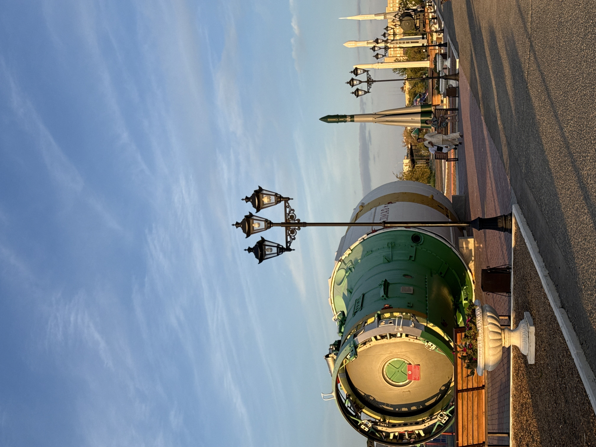 In Baikonur, Kazakhstan, a street scene unfolds with a vast, decommissioned rocket booster as the central exhibit. The booster, painted in prominent green and white, lies horizontally on the ground. To its right, a smaller rocket stands vertically, hinting at the area's cosmodrome history. The sky above is a soft blue, streaked with wispy white clouds. Several ornate, black lampposts with three lamps each punctuate the foreground and midground, casting long shadows across the paved walkway. Benches are placed along the walkway, one occupied by two women, one of whom is looking away from the camera. A large, white planter filled with blooming flowers sits near the rocket booster. The overall atmosphere is one of historical commemoration and public space, bathed in the warm glow of late afternoon or early evening sunlight.