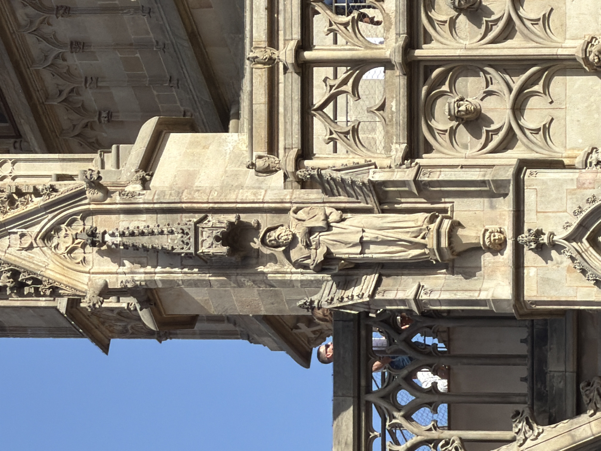 This is a close-up, eye-level, outdoor, daytime shot of the exterior facade of a gothic style building, likely a cathedral or church, in Barcelona, Spain. The facade is adorned with intricate stone carvings, including gargoyles, decorative tracery, and statues. In the foreground, a large stone statue of a robed figure with a hood and beard stands prominently. To the left, a stone balustrade with gothic arched openings runs across the frame. Behind the balustrade, several people are visible, looking out from a higher level of the building. One man with sunglasses is clearly visible in profile, looking to the left. The sky is a clear blue. The architecture suggests a historical and religious context.