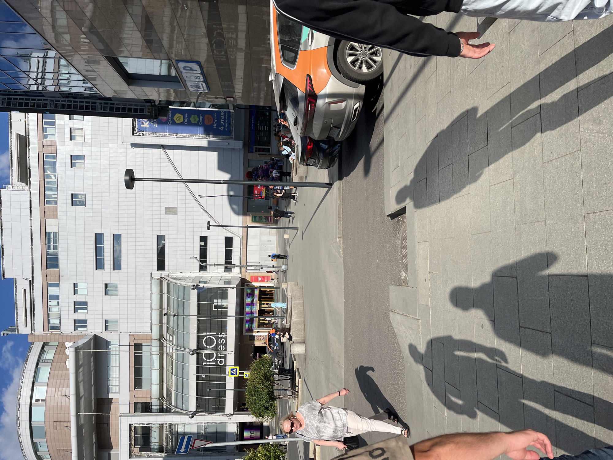 This media file captures a sunny daytime scene on a city street in Moscow, Russia. The foreground features the long shadows of several people cast on a grey paved sidewalk. A person's arm, wearing a black sleeve and a bracelet, is visible in the bottom right corner, suggesting they are the photographer or are interacting with the scene.
In the midground, a street scene unfolds with several cars parked on the side of the road. One car, an orange and silver SUV, is prominently featured, appearing to be involved in a minor fender-bender with a dark car behind it. People are milling about near the vehicles, some appearing to be onlookers or involved parties.
The background is dominated by modern multi-story buildings with glass facades and reflective surfaces. A large banner with Cyrillic text is visible on one of the buildings, advertising "Clothing, Footwear, Sports, Toys" across four floors at "ATRIUM RIM". Another sign reads "1001 dress". Street signs, including a blue "P" indicating parking and traffic signs, are also present. The overall impression is a bustling urban environment on a clear, bright day.