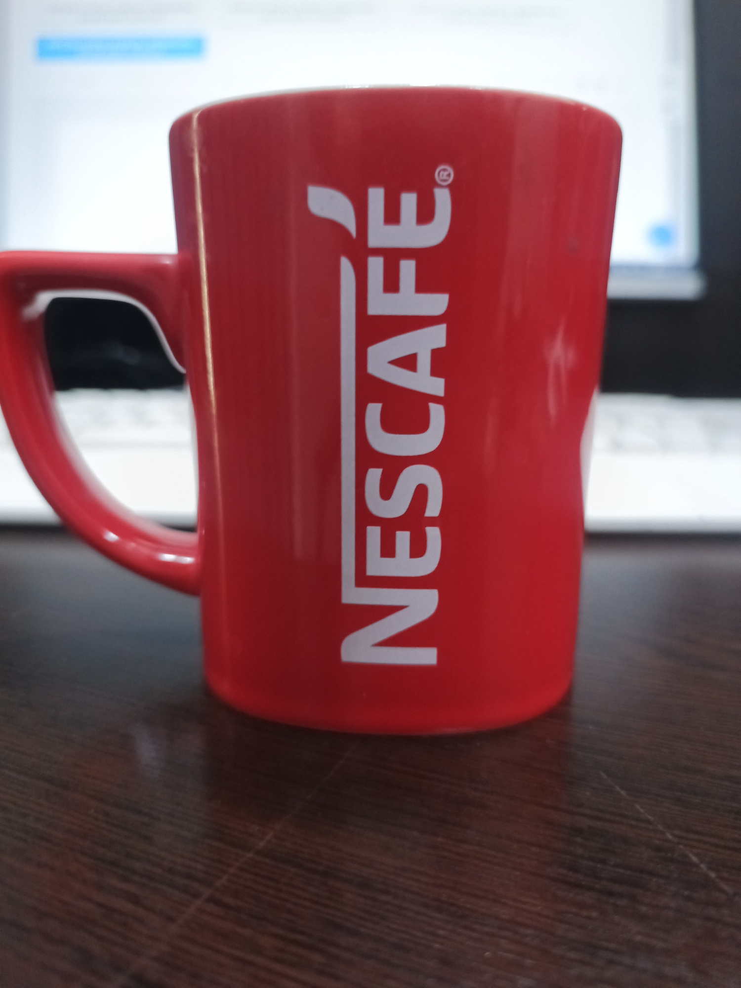 A close-up, low-angle shot shows a red Nescafe mug on a dark wooden surface. The mug is cylindrical with a handle on the left side. The Nescafe logo, in white, is printed vertically on the front of the mug. In the blurred background, a laptop screen is visible, displaying a light blue bar at the top. The scene appears to be indoors, on a desk. The location is identified as Qorowul, Uzbekistan.