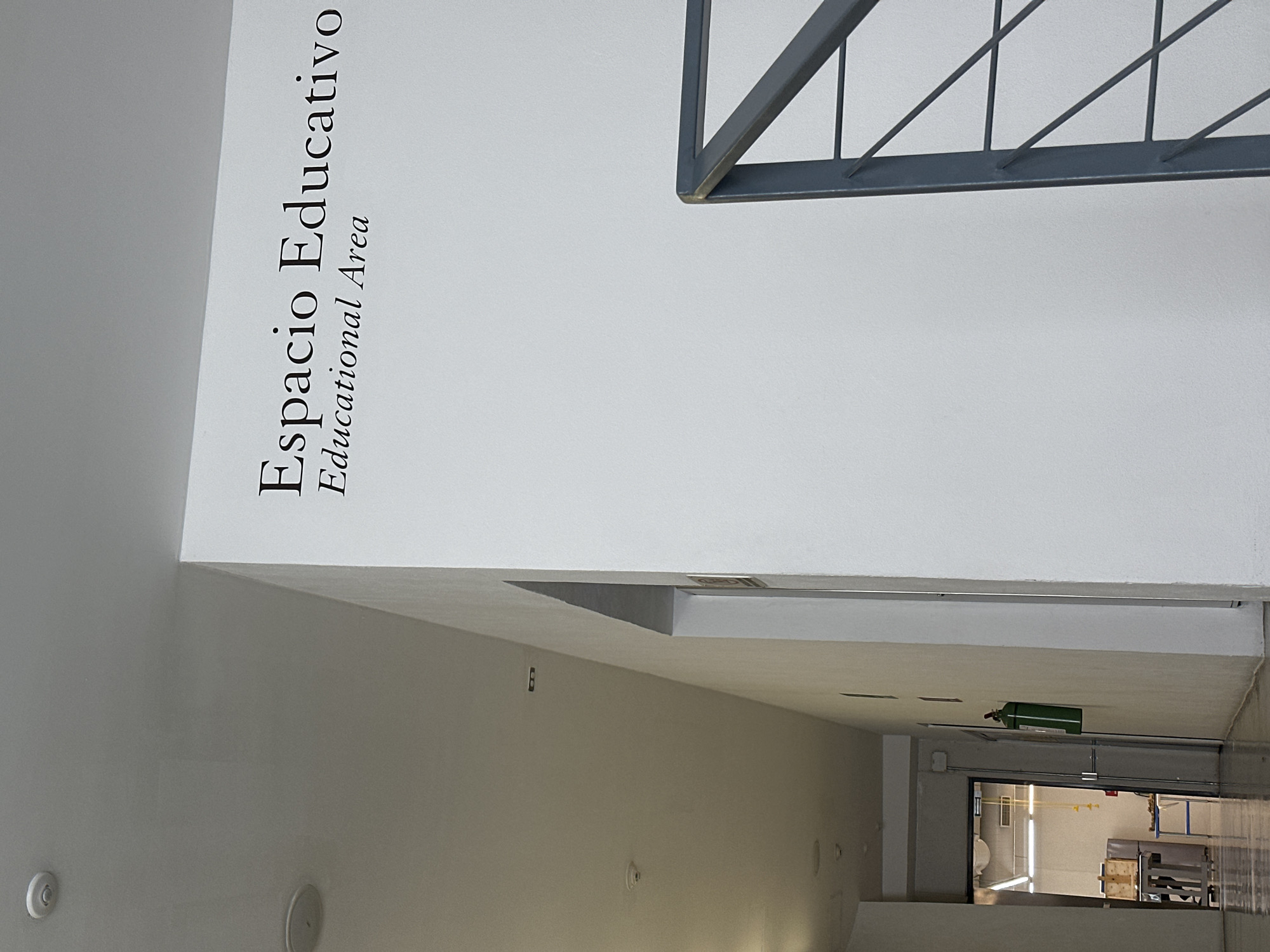 This image captures the interior of a modern building, likely an educational or institutional facility, in San Bernardino Tlaxcalancingo, Mexico. The perspective is angled, looking down a corridor or open space from a slightly elevated vantage point.
The dominant features are clean, light-colored walls, predominantly white. On a prominent white wall in the upper left, the text "Espacio Educativo" (Educational Space) and, directly below it, "Educational Area" are printed in black, indicating the function of this part of the building.
In the upper right quadrant, a dark gray metal railing or structural element is visible, featuring diagonal and horizontal bars, suggesting a staircase, balcony, or walkway. The walls extend downwards into a hallway or a series of rooms. Along the left wall in the mid-ground, a circular white fixture, possibly a speaker or smoke detector, is visible. Further down the corridor, mounted on a wall or ceiling, is a green cylindrical object, which appears to be a fire extinguisher.
At the far end of the visible space, an open doorway or glass partition leads into another room. This room contains some industrial-looking equipment, including what appears to be pallets or storage structures, and possibly a sink or toilet in the deeper background, suggesting a utility area, workshop, or storage space. The area is well-lit by artificial indoor lighting. No people are present, lending a quiet, unoccupied atmosphere to the scene. The overall impression is one of functionality and modern design within an educational context.