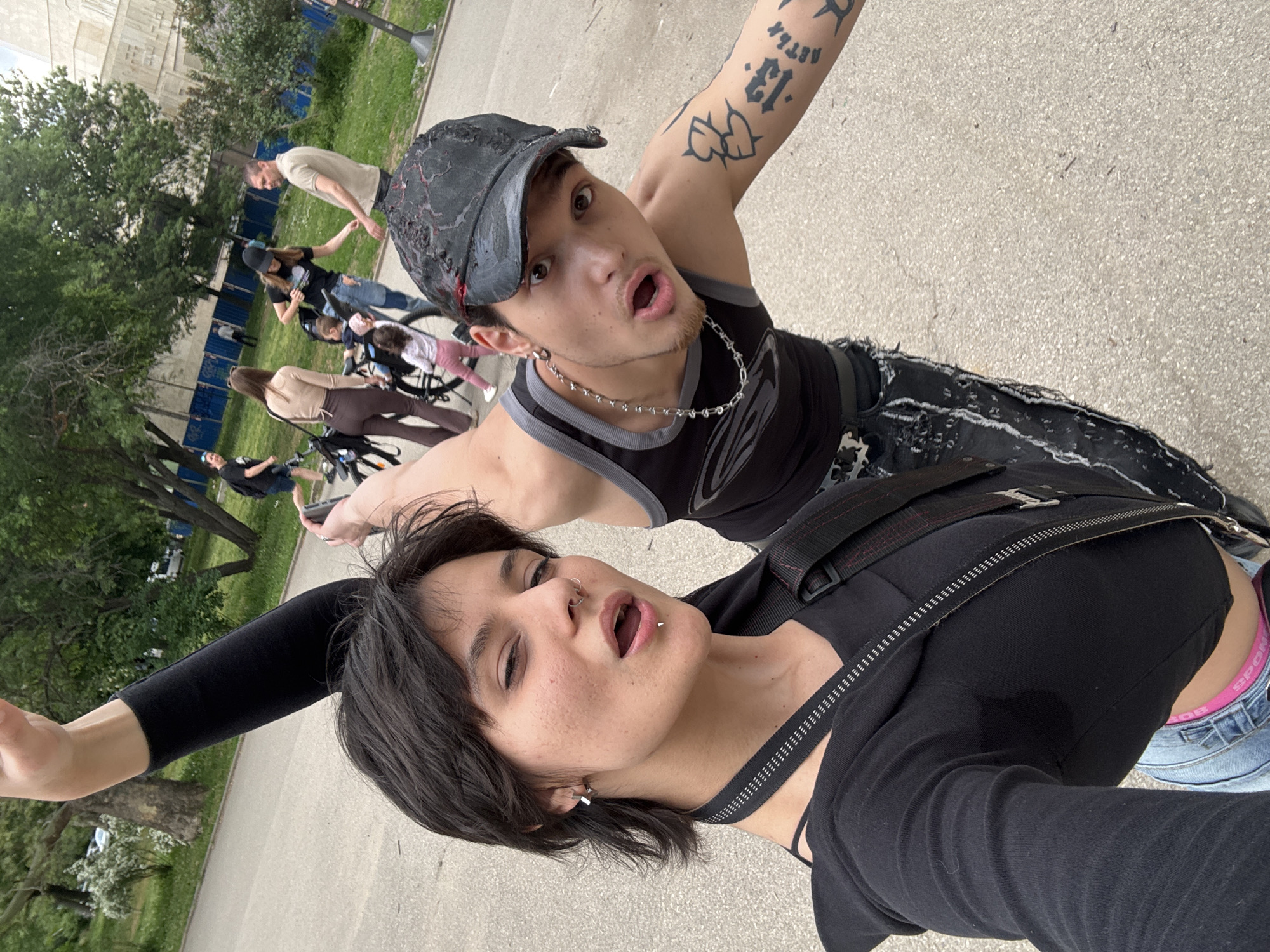 Two young people, one wearing a black tank top, ripped jeans, a cap, and a silver chain and the other wearing a black top with a harness are taking a selfie. The first person has tattoos on their arm. Behind them are multiple people walking and riding bicycles. Trees and a building are in the background. The picture appears to be taken in a public outdoor space in Sofia, Bulgaria.