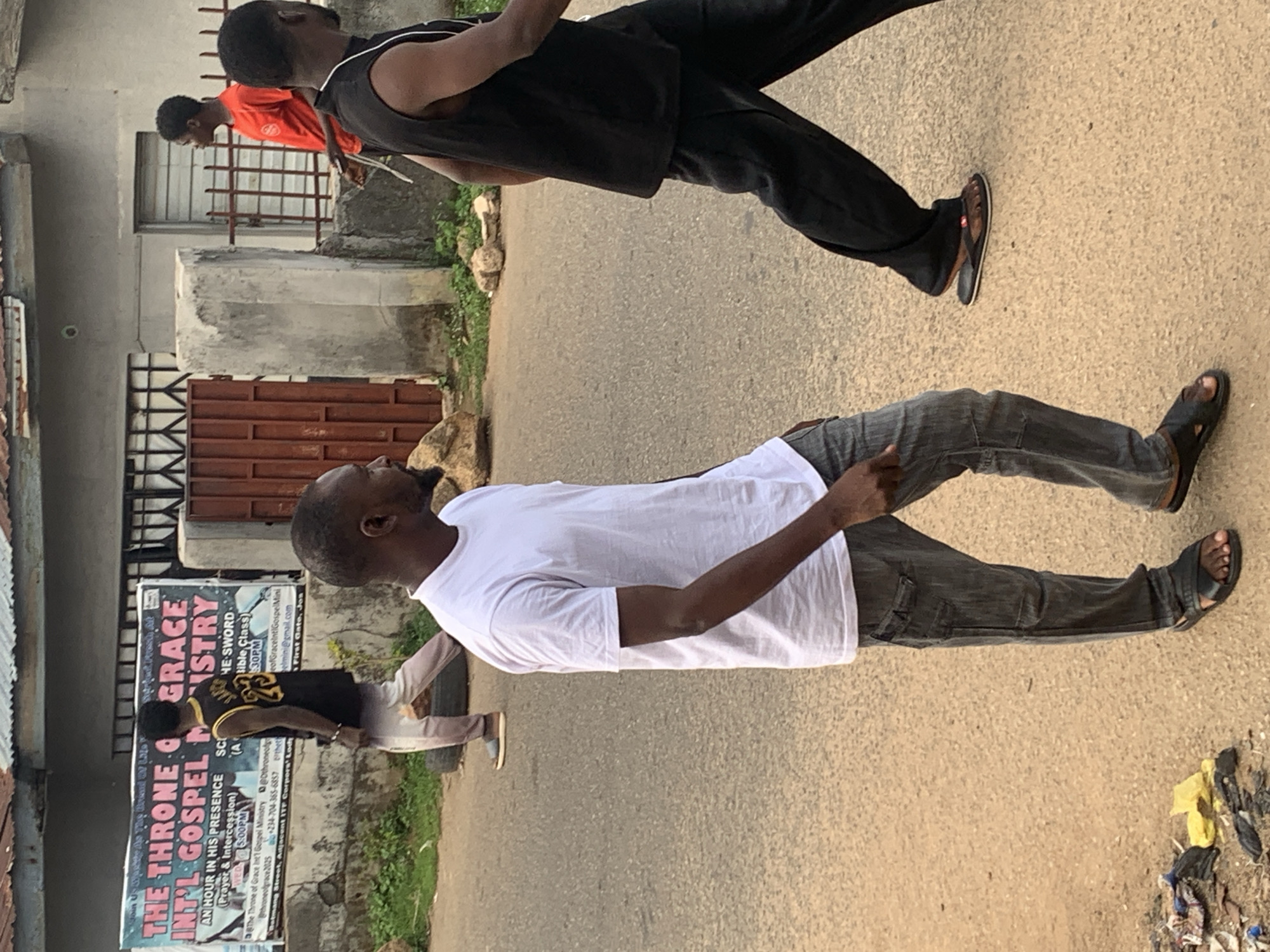 Three males are present, walking down a paved road. One is wearing a white shirt and dark pants, another is wearing a black tank top and dark pants, and the third is wearing a red shirt. A sign for "The Throne Grace Int'l Gospel Ministry" is visible near the road. The location is determined to be Barakin Akawo, Nigeria.