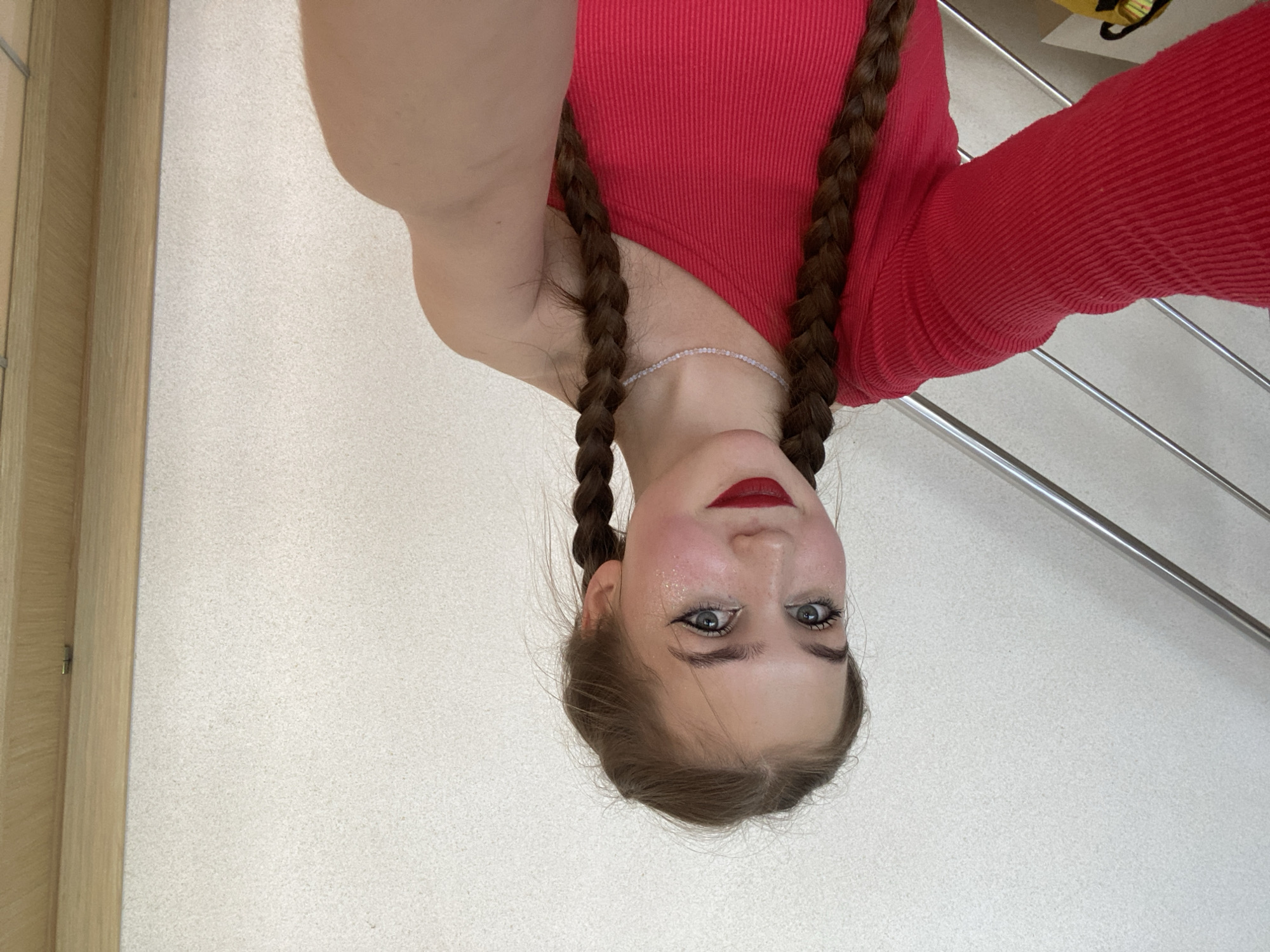 A young woman with long, brown braided hair and blue eyes is taking a selfie. She is wearing a red, one-shoulder top and a delicate pearl necklace. Her makeup includes winged eyeliner, blush, and bold red lipstick. There are subtle flecks of glitter on her cheekbones. She is standing indoors in front of a textured, light-colored wall. To her left, a silver railing is visible, suggesting she might be on a staircase. To her right, a wooden door frame is present. The location within the city of Прикамский, Russia is not discernible from the image alone.