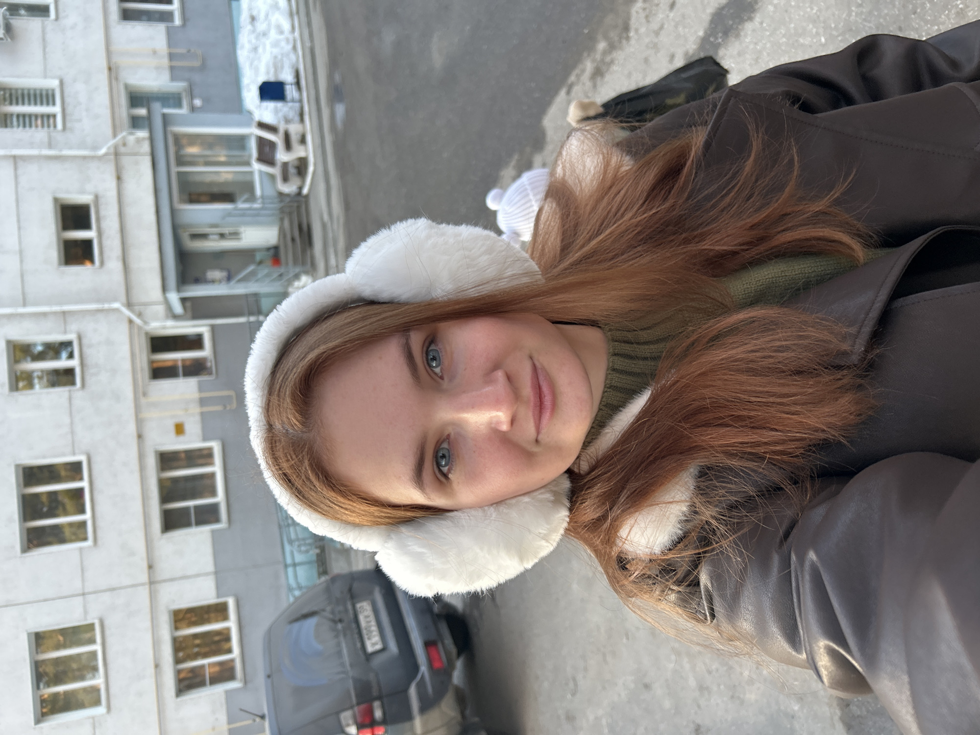 The image shows a young woman in a brown jacket and white earmuffs, taking a selfie in an outdoor setting. She has fair skin, blue eyes, and reddish-brown hair. She's wearing a green knitted sweater beneath the jacket.
In the background is a light gray building with multiple rectangular windows. A dark gray car is partially visible in the lower left corner of the image. Based on visual identifiers such as license plate styling and architectural design, the location appears to be Chaikovsky, Russia.